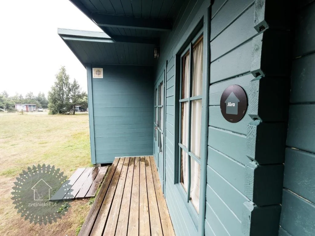 Exterior view of a blue wooden house with a wooden porch, a window with curtains, and a sign with an upward arrow and the word "Jura".