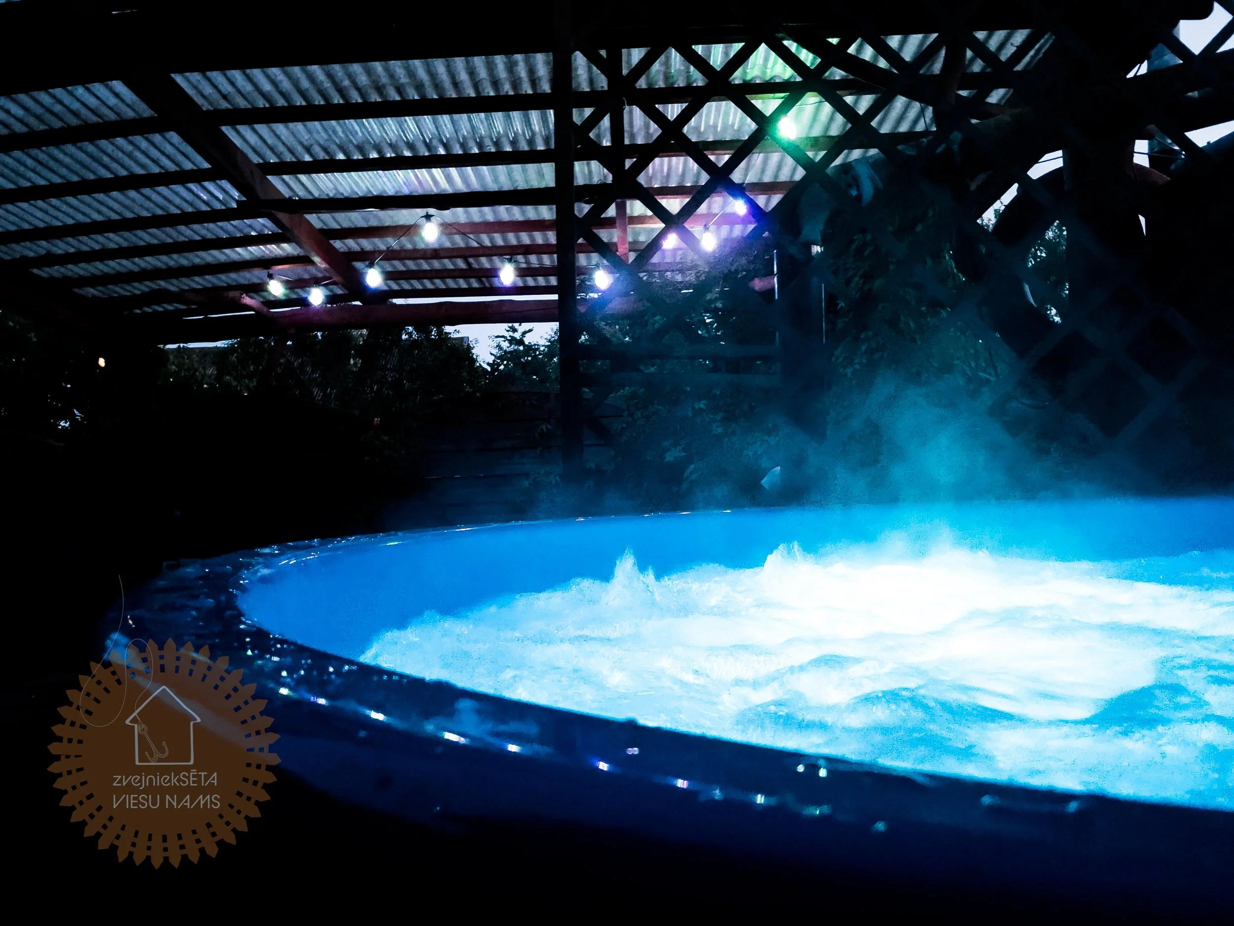 Hot tub bubbling with steam in an outdoor setting, with a wooden roof and string lights overhead during the evening.