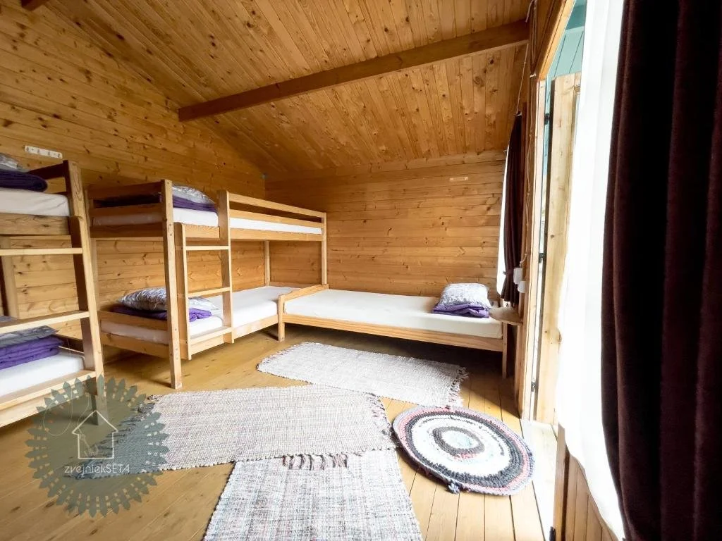 A cozy wooden bedroom with three beds: a bunk bed on the left and a single bed on the right, with a window and dark curtains on the right side.