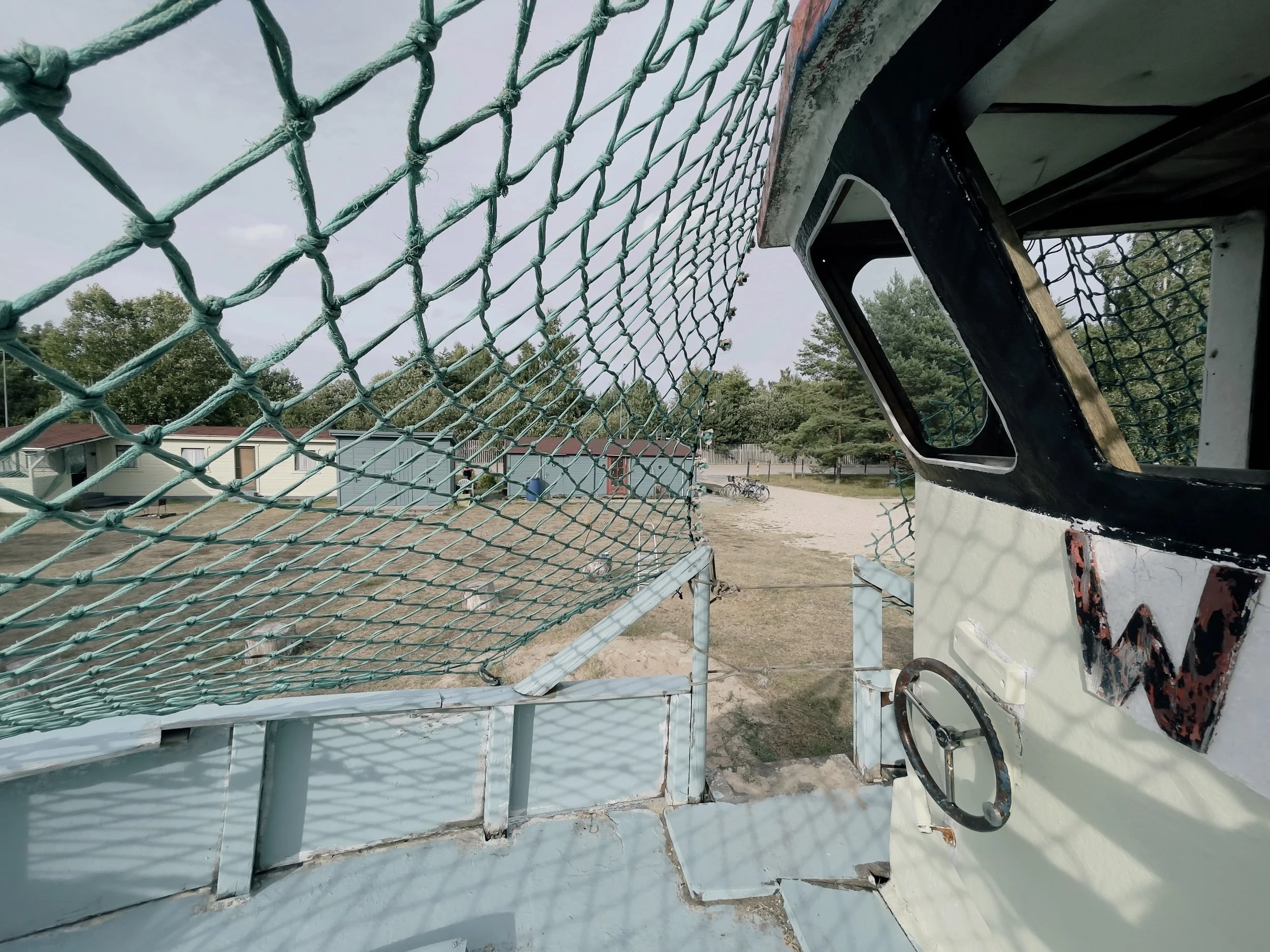 View from inside an old, weathered amusement park ride cabin with a large front window, surrounded by a safety net and metal railing, with outdoor trees and small buildings in the background.