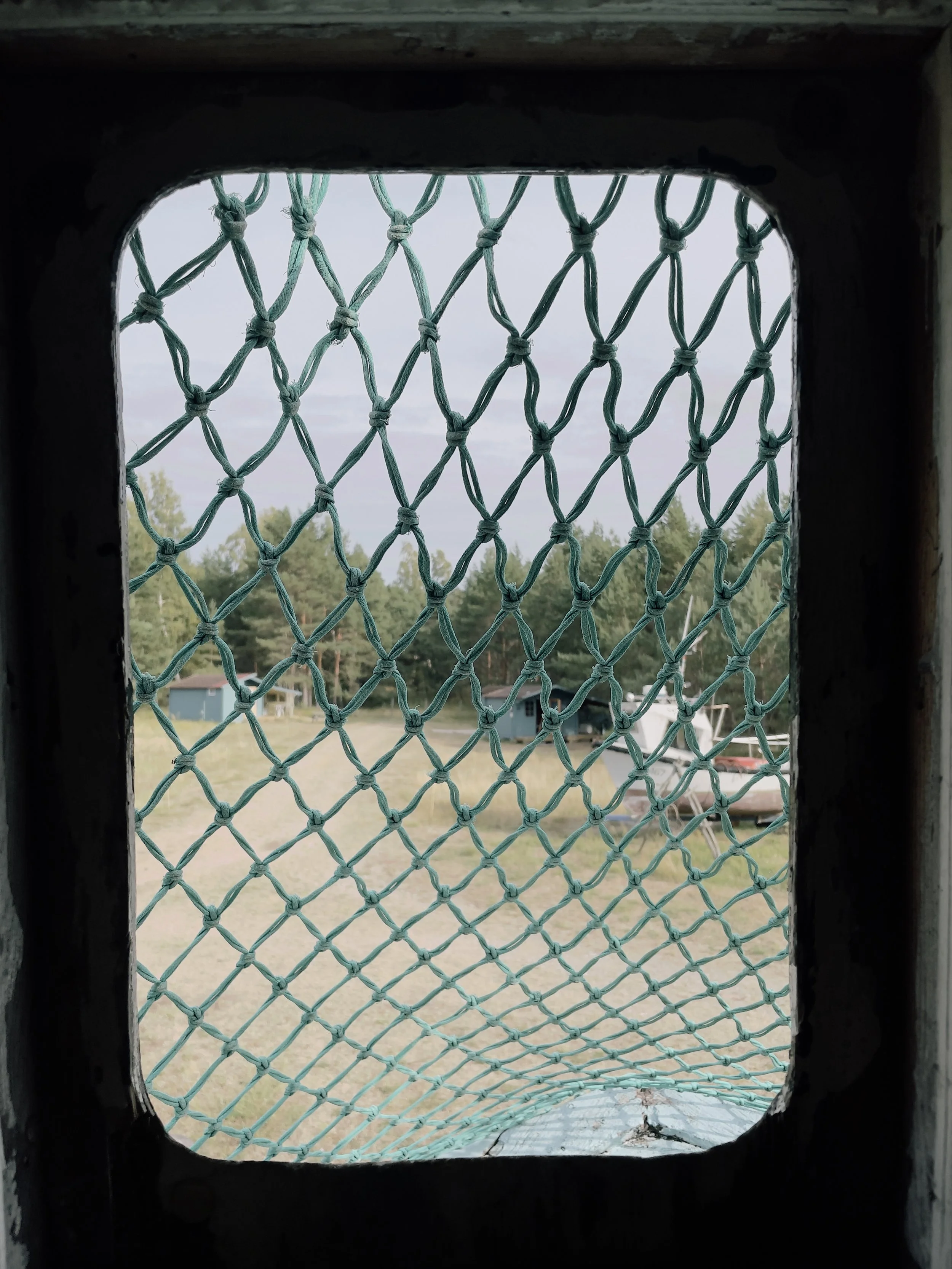 View through a small window with a wire mesh screen, overlooking a grassy yard with trees, a small building, and a docked boat in the distance.