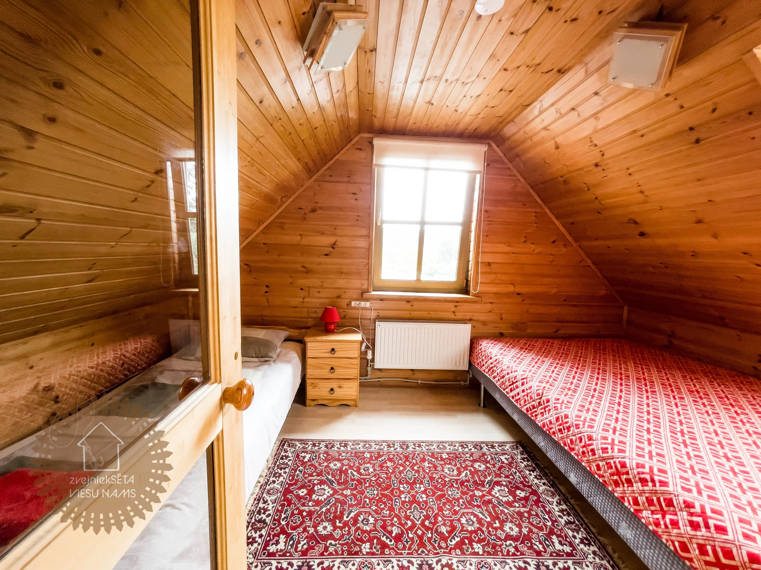 A cozy attic bedroom with wooden paneled walls and ceiling, featuring a double bed with a red patterned bedspread, a single bed with a white sheet, a small wooden nightstand with a red lamp, a window with white blinds, a radiator beneath the window,