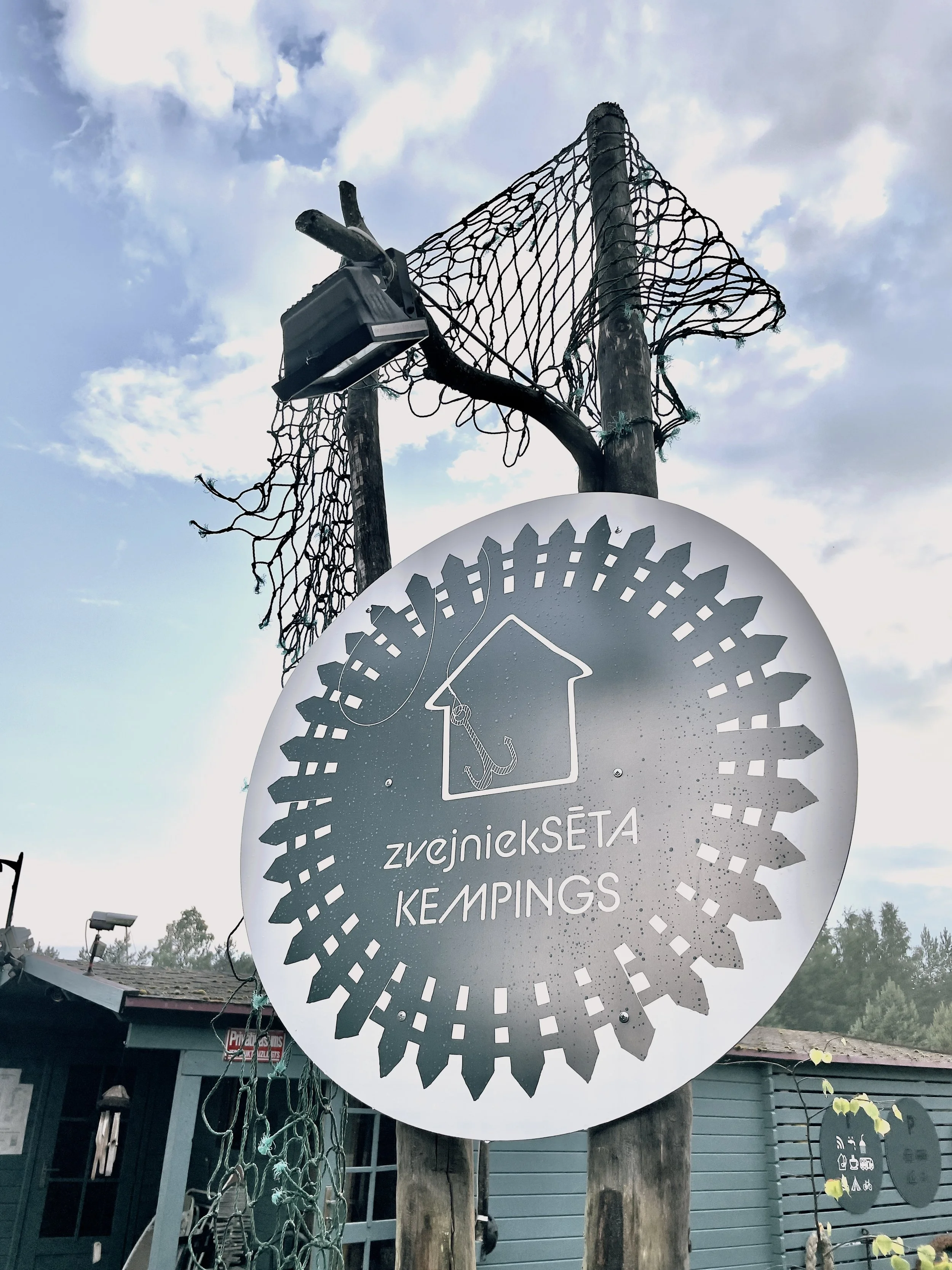 Sign for ZvejnickSeta Kempings with a house and fishing hook logo, mounted on a wooden post against a cloudy sky.