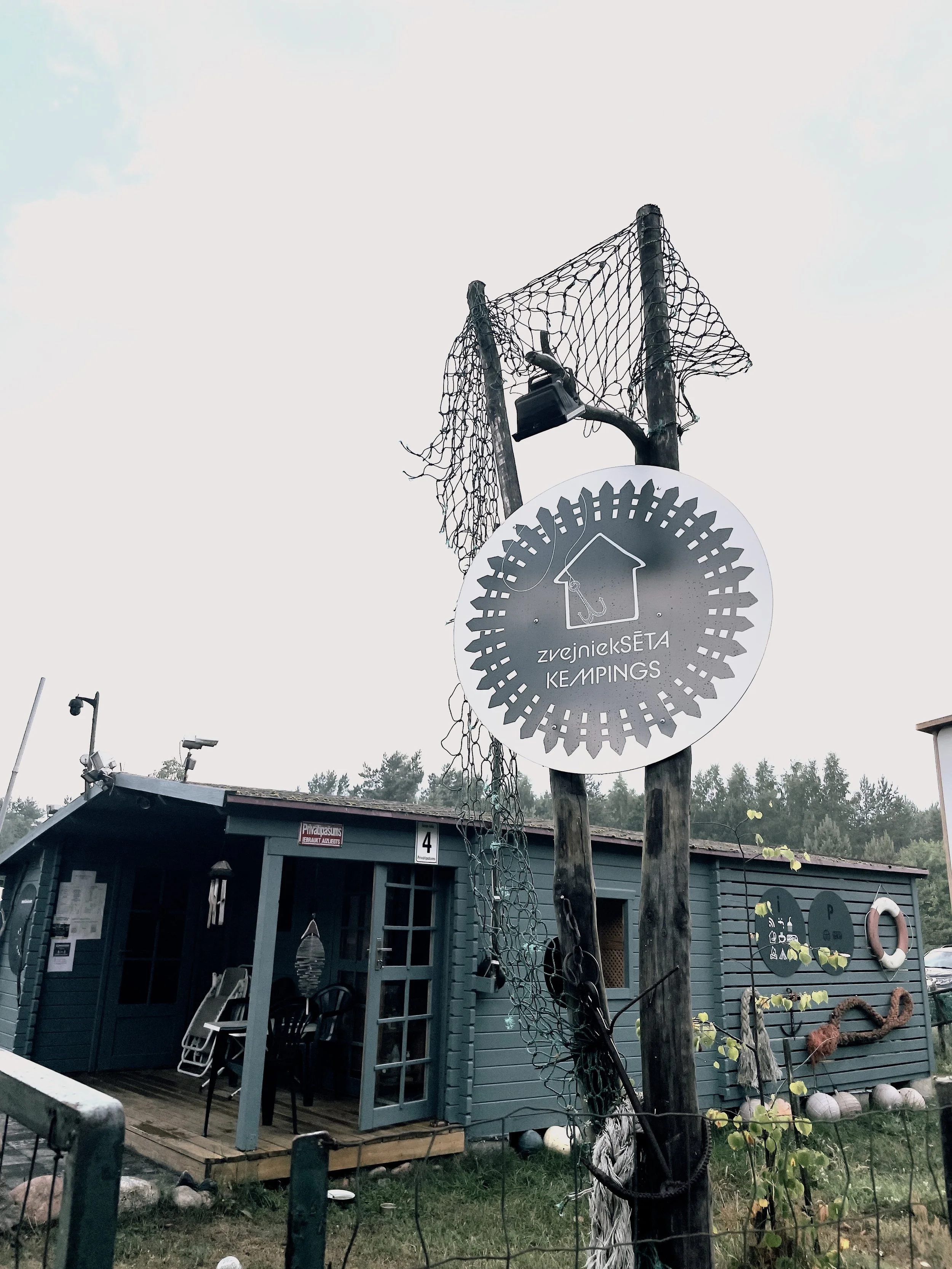 A blue wooden cabin with an open door, surrounded by outdoor chairs and decorations. A sign on a wooden pole reads 'ZvejnickSETA KEMPINGS' with an house and fish hook icon, and a tattered fishing net is draped over the pole.