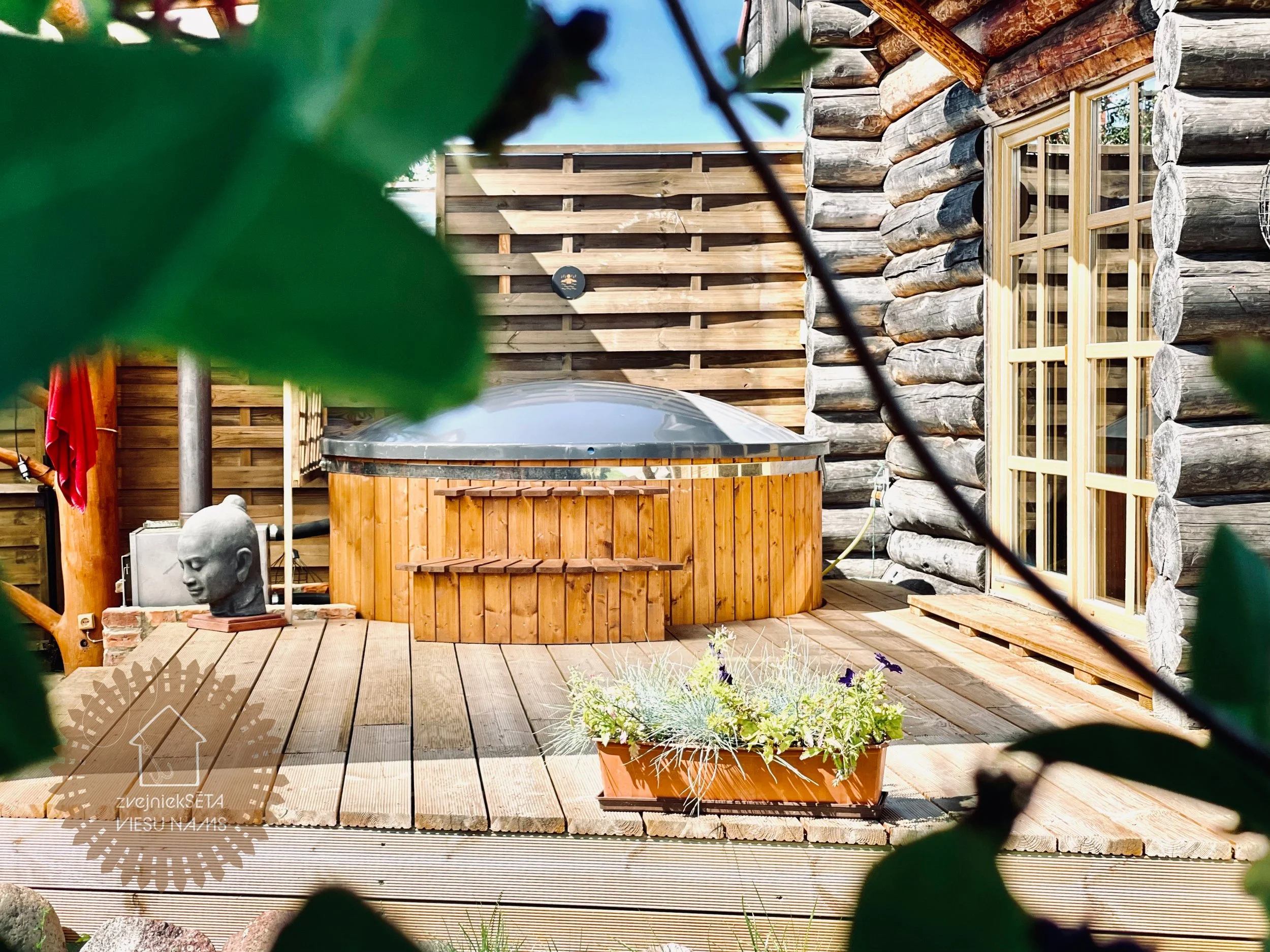 Outdoor wooden deck next to a log cabin with a hot tub, a plant box, a stone bust sculpture, and leafy branches partially obscuring the view.