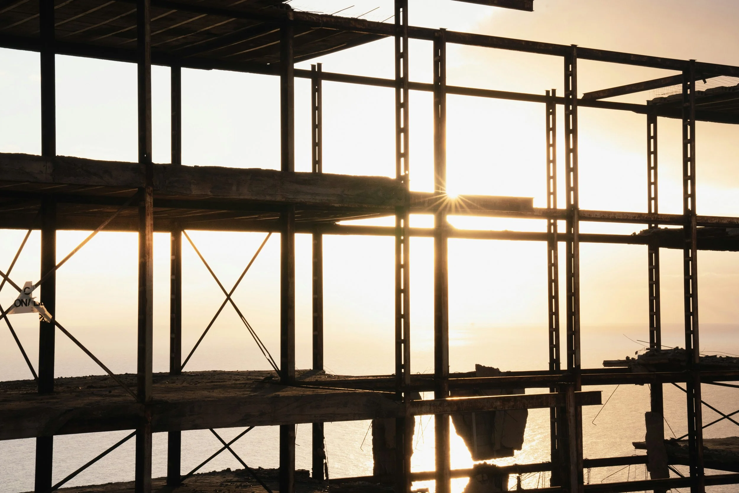 Silhouette of a building under construction with the sunset in the background, showing structural beams and scaffolding.