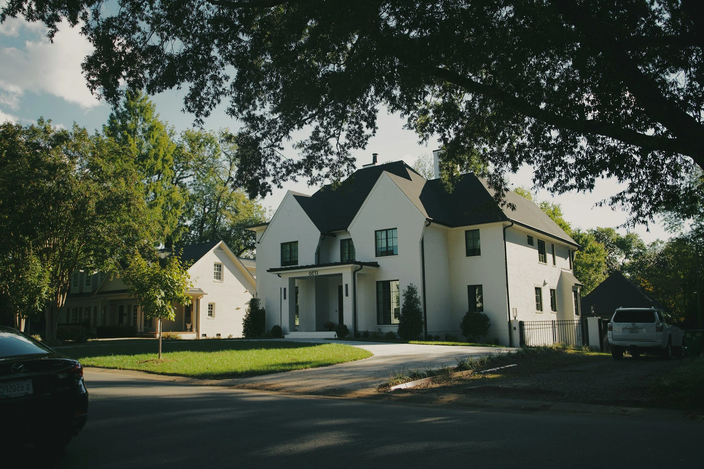 A white two-story house with black roof and window frames, surrounded by trees and parked cars in a suburban neighborhood.