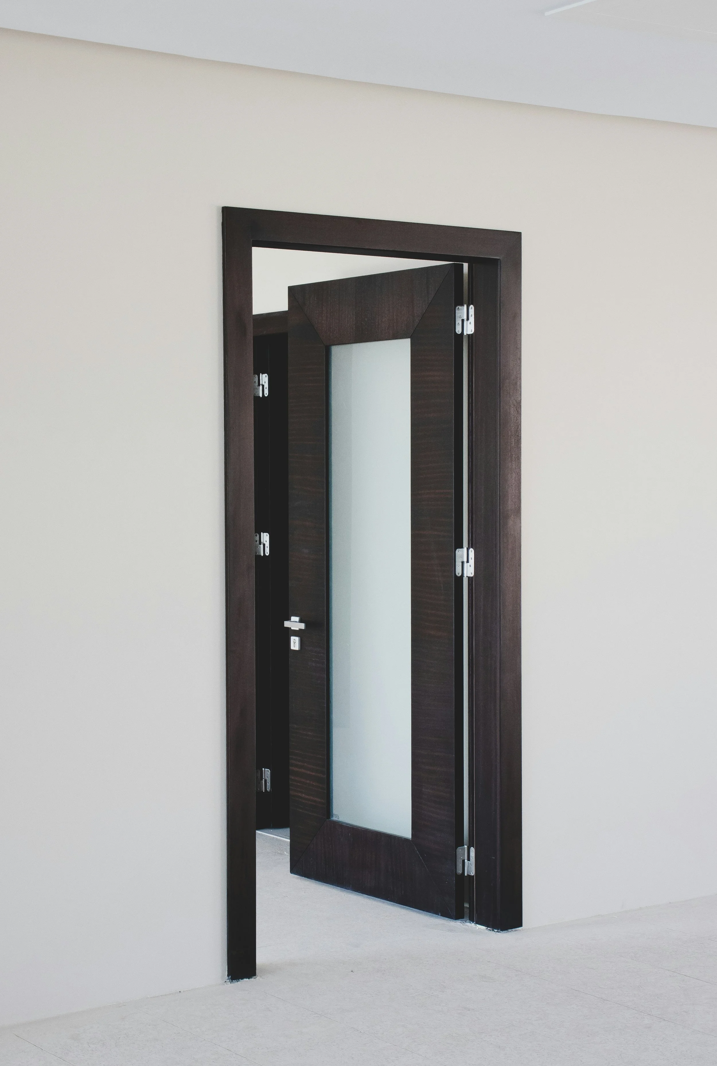 An open dark wooden door with a frosted glass panel, door handle, and hinges, leading into a room with plain white walls and a light-colored carpeted floor.