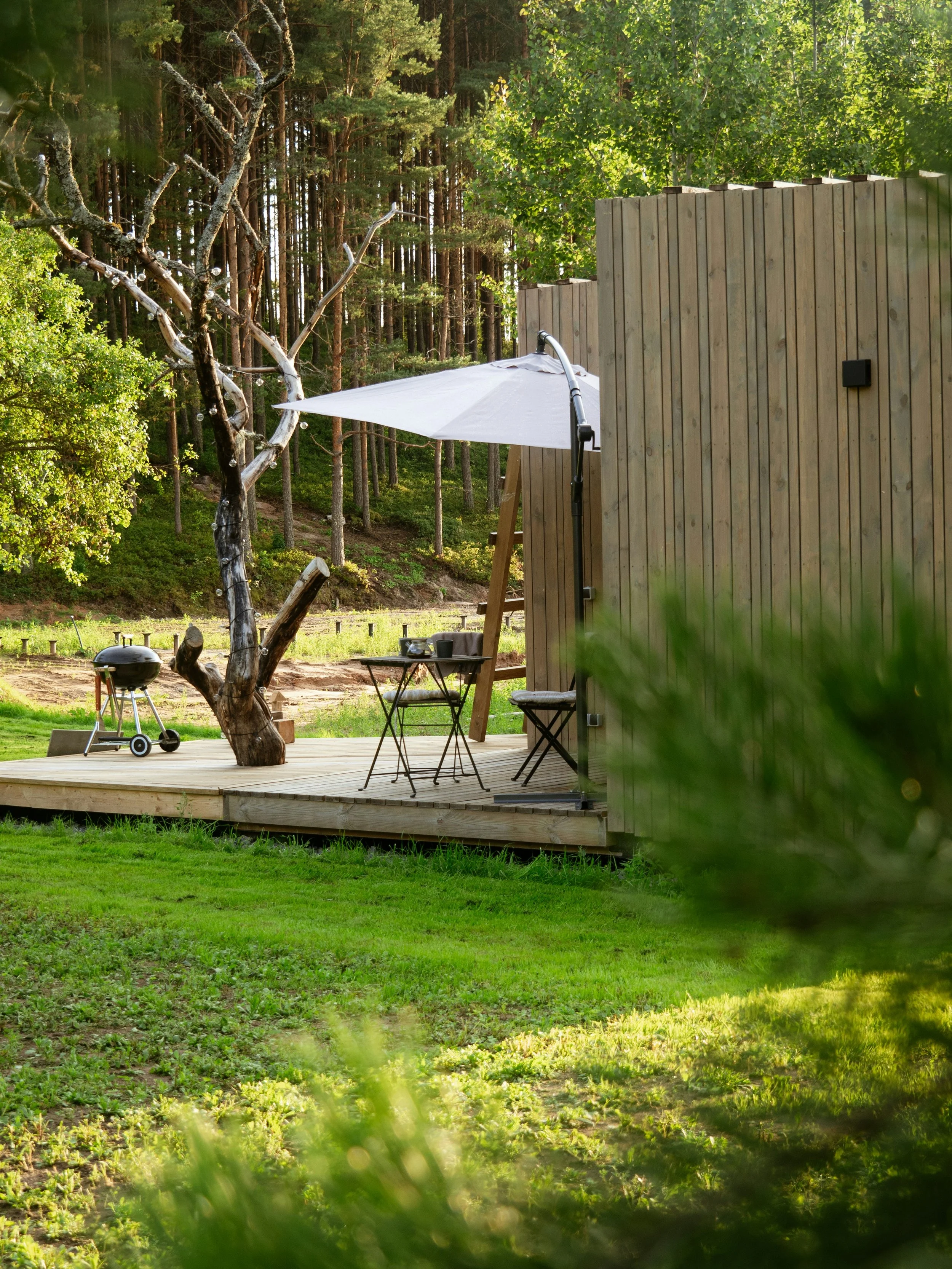 A wooden deck in a backyard with a small round table, two chairs, a white patio umbrella, and a barbecue grill, surrounded by green grass, trees, and a wooden privacy fence.