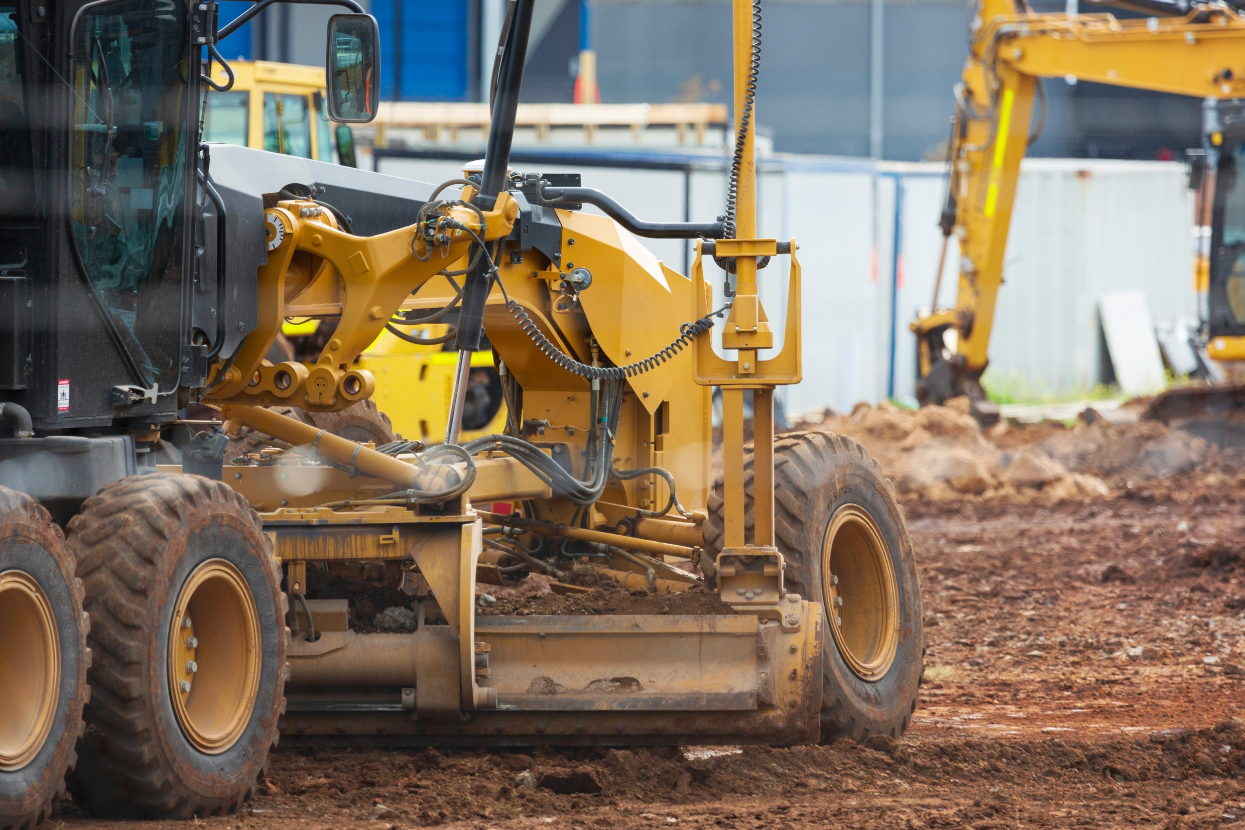 Construction site with yellow and black heavy machinery, including a skid steer loader in the foreground and excavators in the background, on a dirt surface.