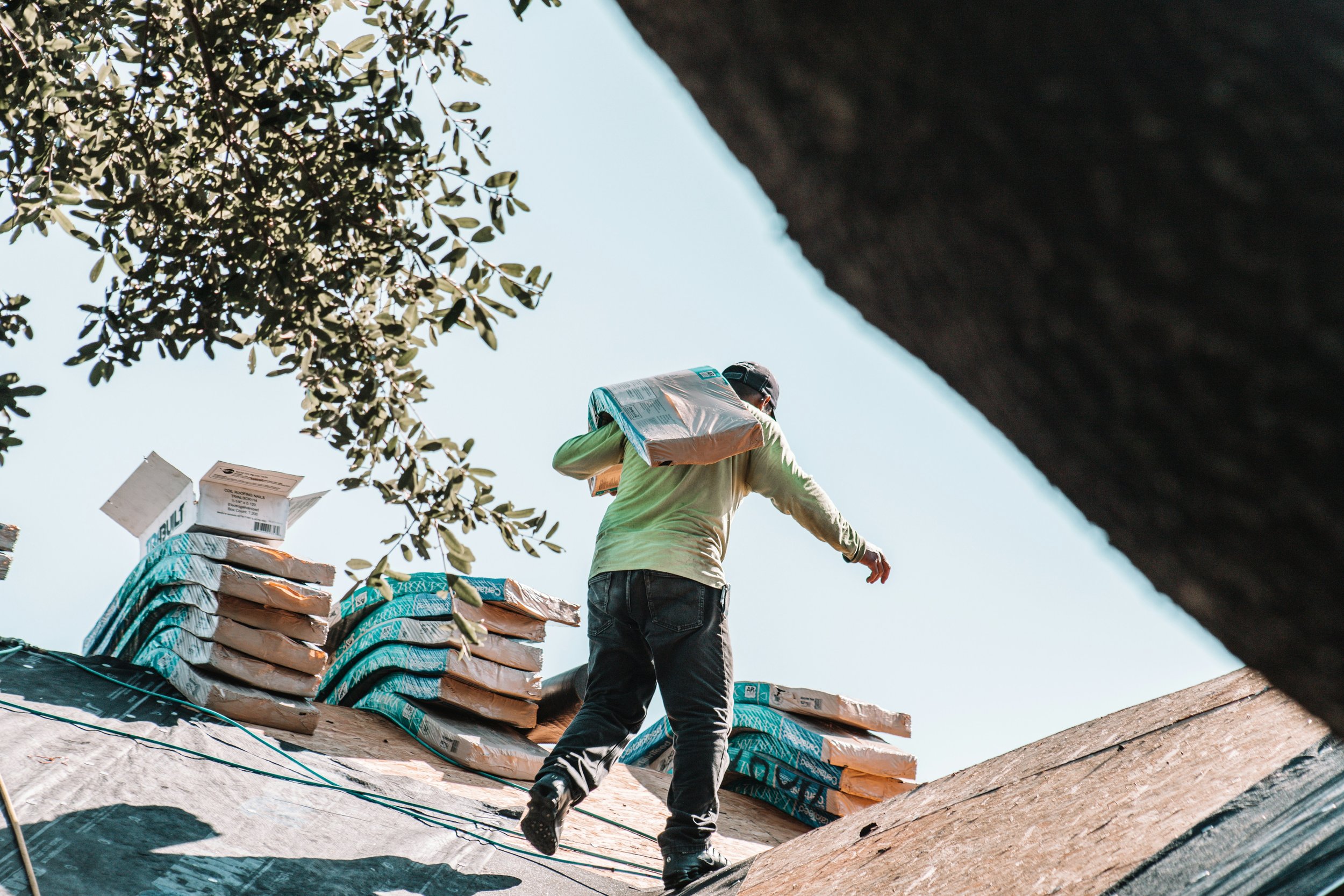 A man carrying a bag of roofing materials walking on a roof under blue sky, with green foliage partially obscuring the view.
