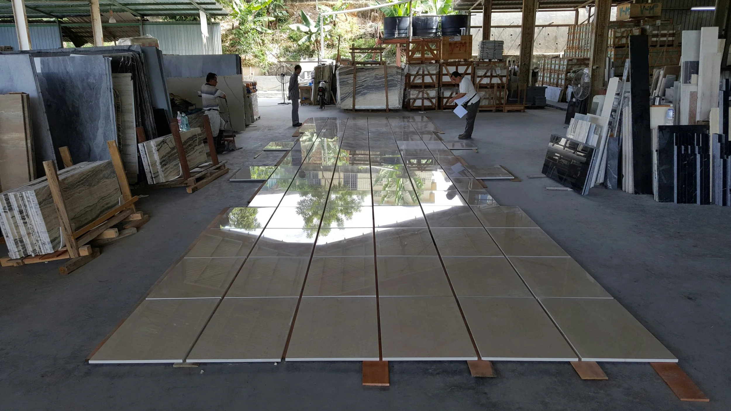 Large polished marble tiles laid out on the floor of a warehouse, with several workers inspecting or preparing them. The warehouse has stacked slabs of marble or stone along the sides and in the background.