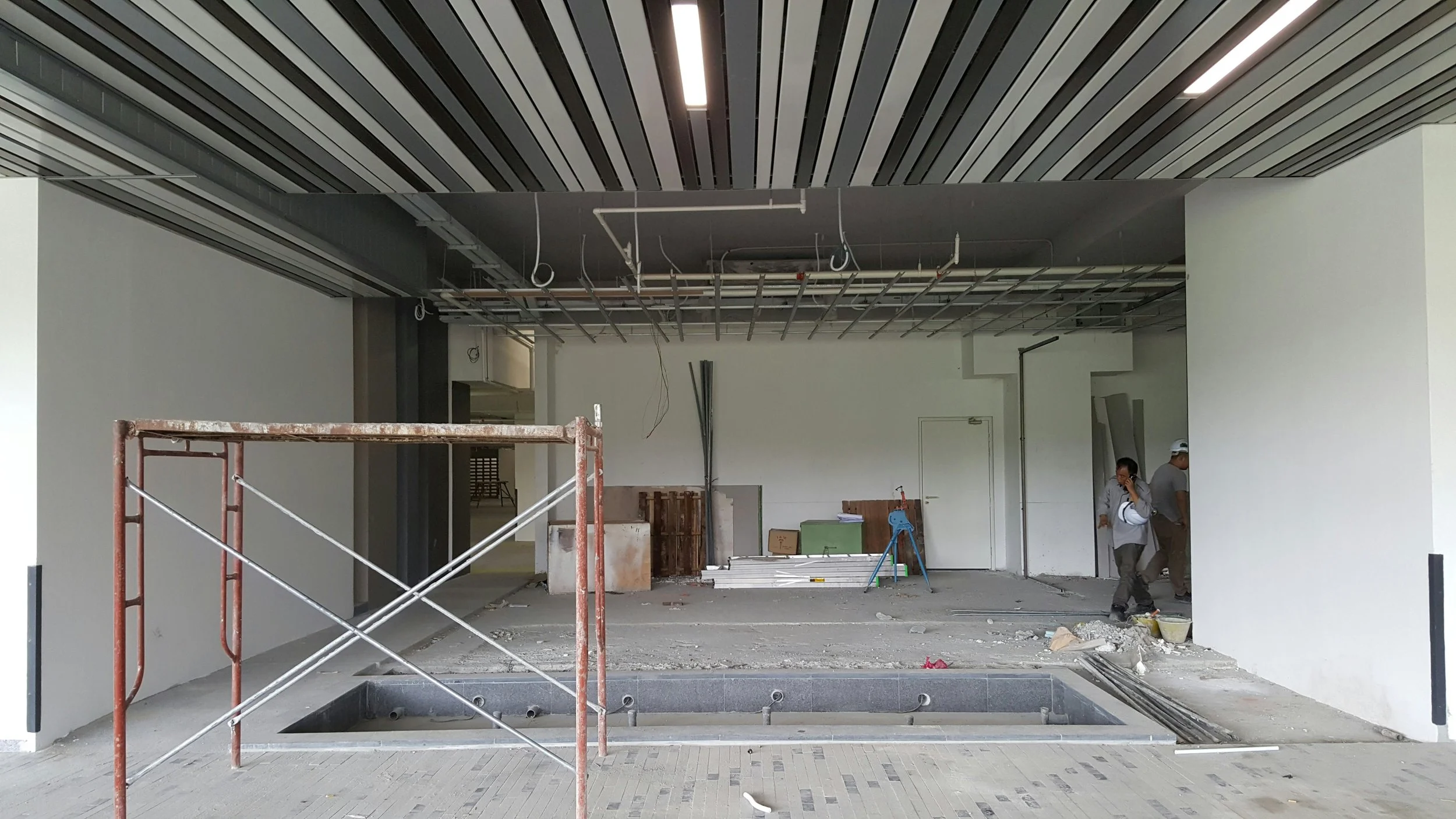 Construction site inside a building with white walls, metal ceiling panels, and construction workers walking. There is scaffolding, construction materials, and exposed wiring overhead.