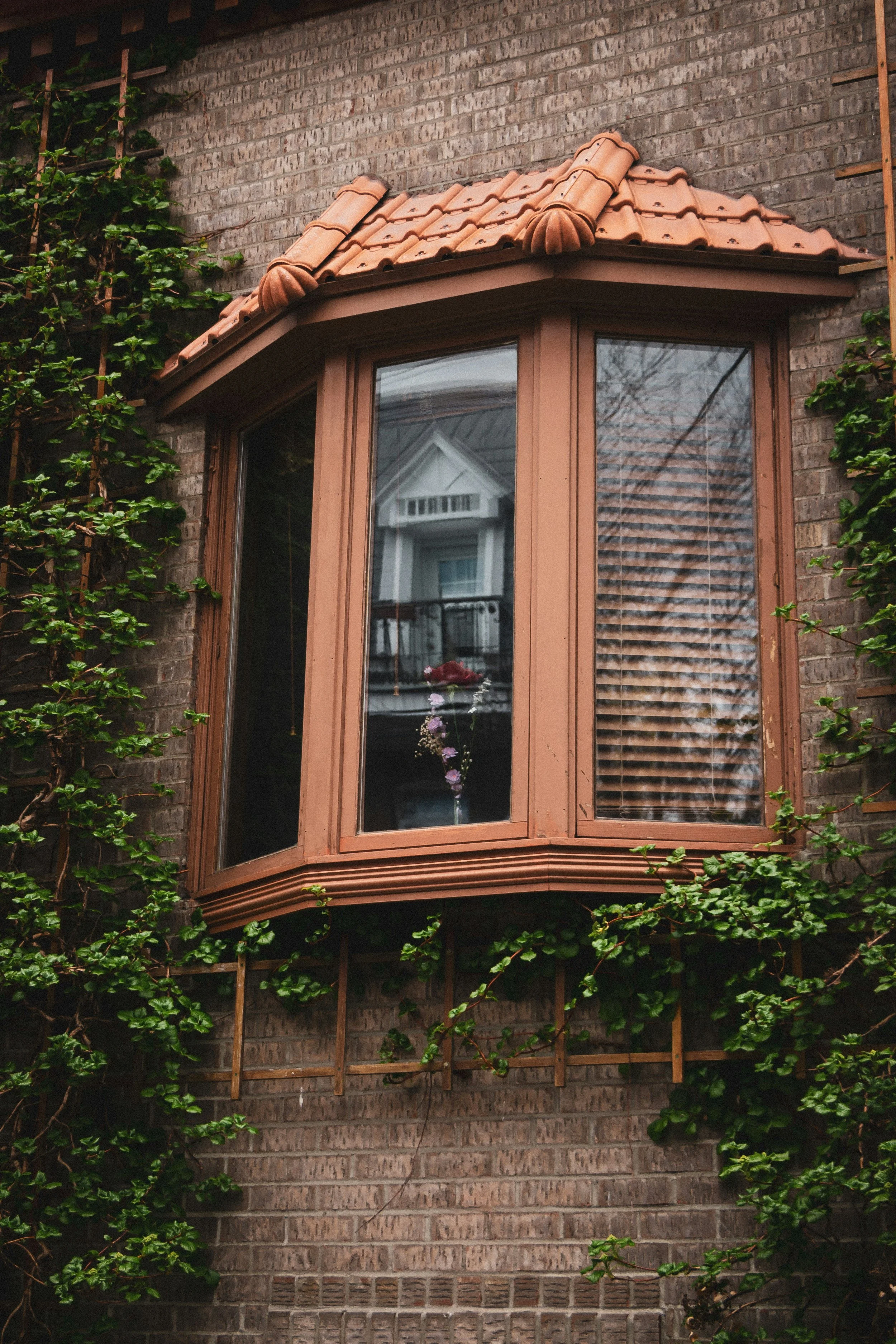 A brick house exterior featuring a flower box window with a terracotta tiled roof, surrounded by green climbing plants.