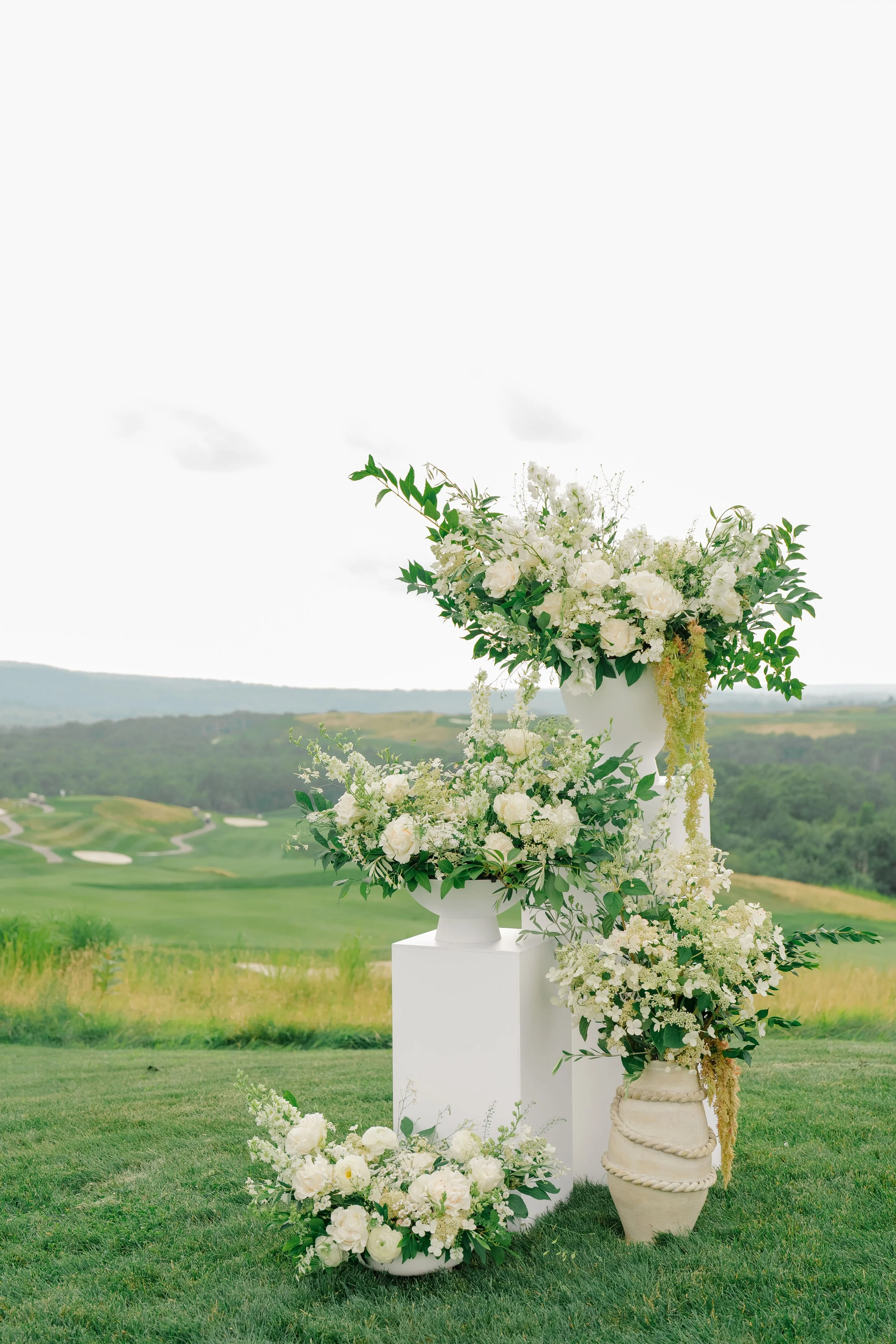 White floral arrangements with greenery on white pedestals and vessels, set outdoors on a grassy area with a golf course in the background.