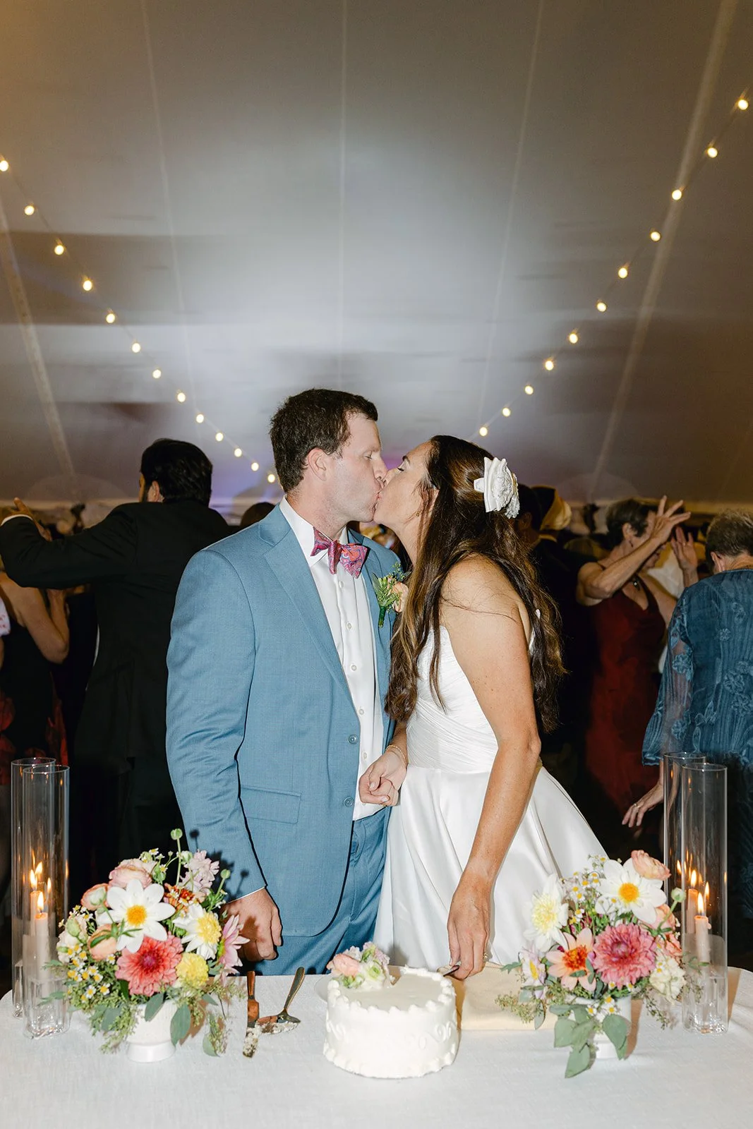A bride and groom kissing at their wedding reception with floral centerpieces and a cake on the table.