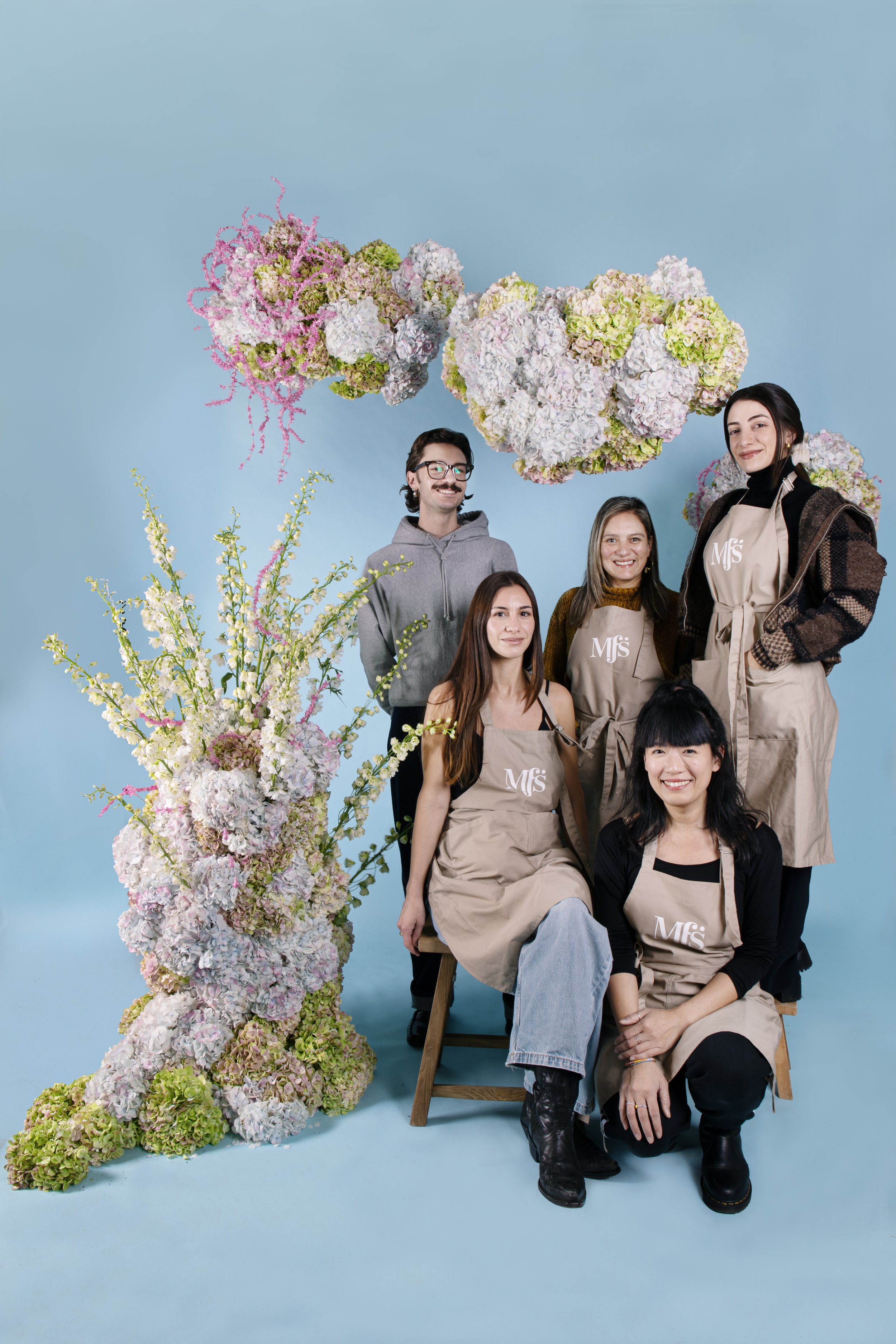 Group of five people posing with large floral arrangements on a light blue background. Four are wearing beige aprons with 'MFS' logo, and one is in casual clothes. The floral arrangements include pastel-colored hydrangeas and other flowers.