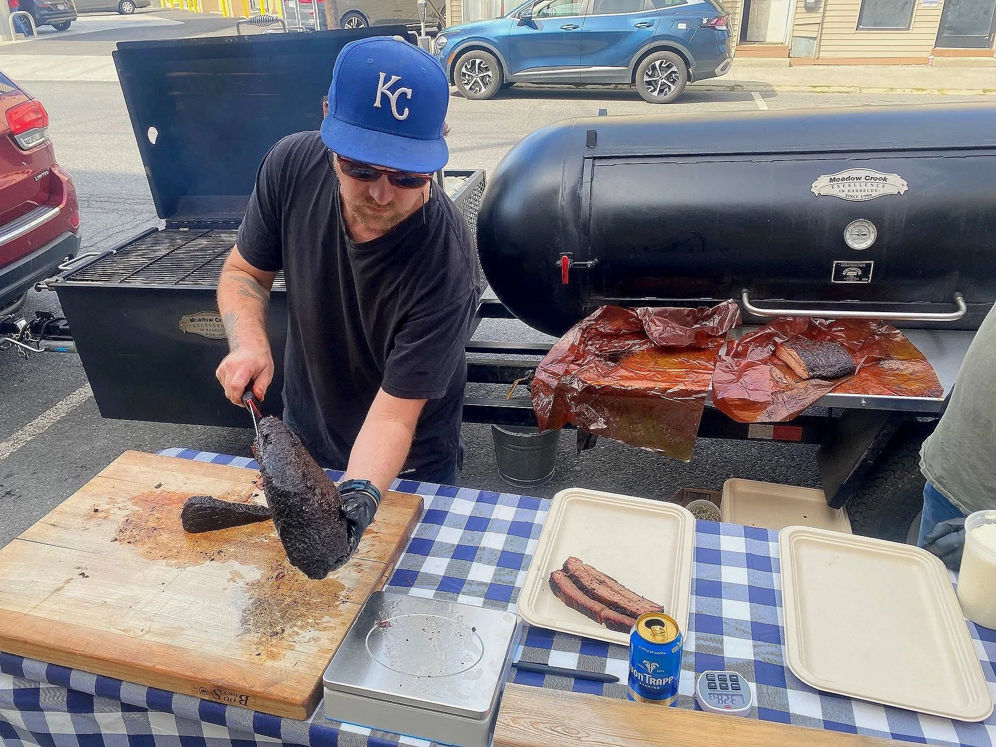 A man with a Kansas City cap, sunglasses, and black t-shirt is slicing smoked meat from a large smoked brisket on a cutting board at an outdoor barbecue stand. There is a black smoker grill behind him, with some smoked paper liners, and a plate with a cut of smoked meat. A can of Pabst Blue Ribbon beer and digital scales are on the checkered tablecloth.
