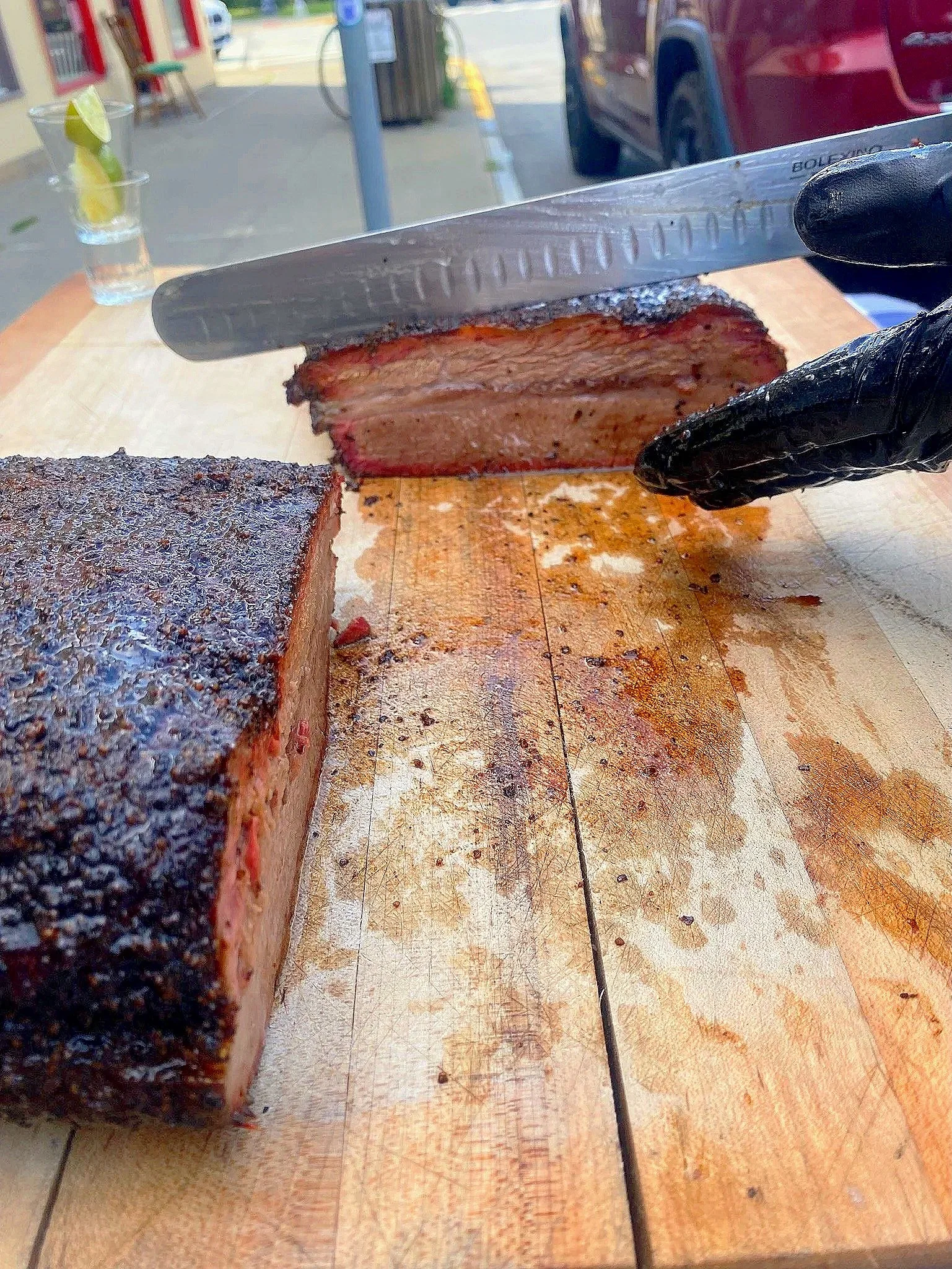 A person slicing a large smoked brisket on a wooden cutting board outdoors, with a knife and a gloved hand visible.
