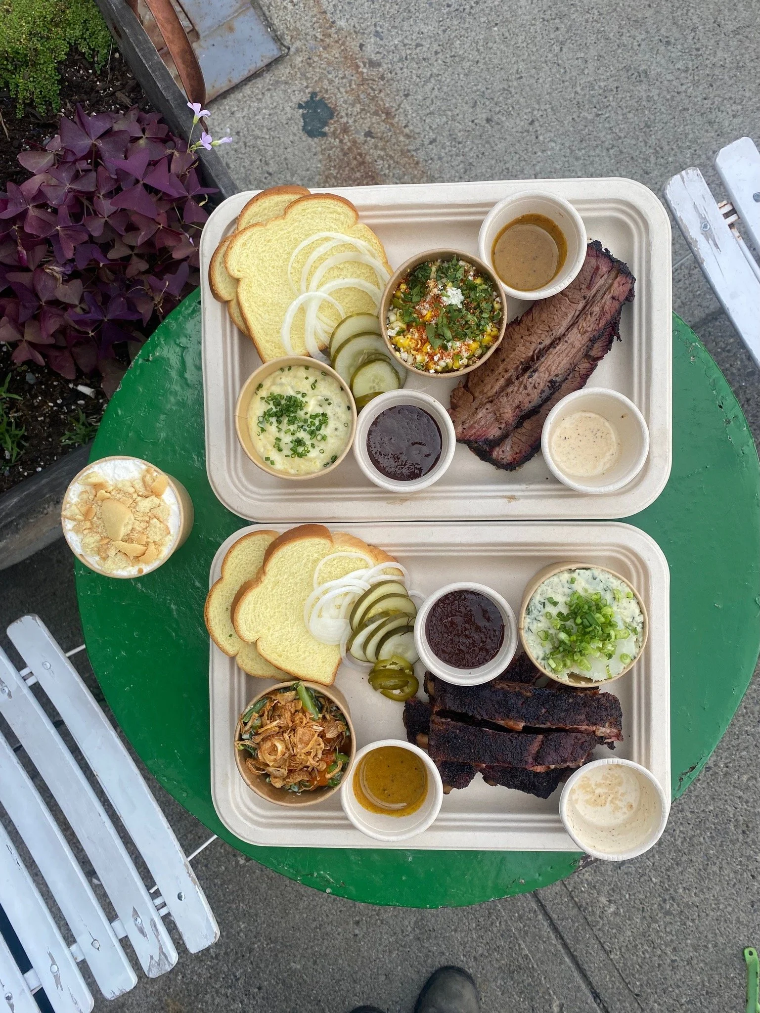 Two trays of barbecue food on a green table, with sides of bread, pickles, onions, jalapenos, and various sauces, along with a small bowl of potato salad and a cup of ice cream.
