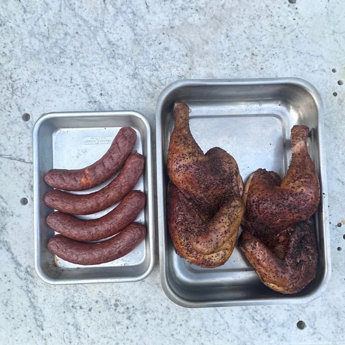 Grilled sausage links on the left and seasoned grilled chicken thighs on the right in metal trays on a concrete surface.