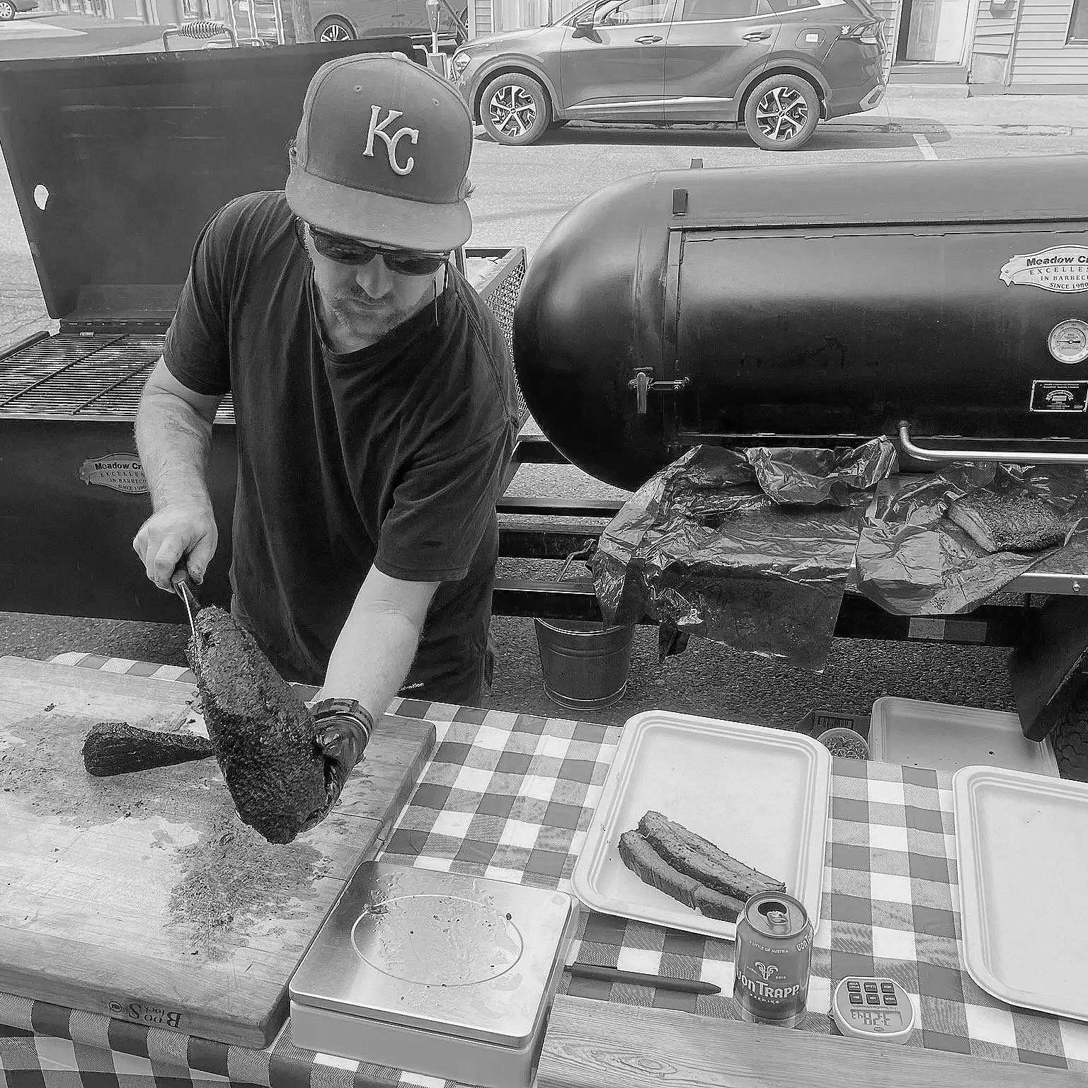 A man wearing sunglasses, a Kansas City cap, and a black t-shirt is slicing a smoked or grilled piece of meat on a wooden cutting board. The scene is set outdoors at a barbecue event with a checkered tablecloth, plastic containers, a can of beer, and a digital scale on the table. There is a large black smoker or grill in the background and a parked car on the street.