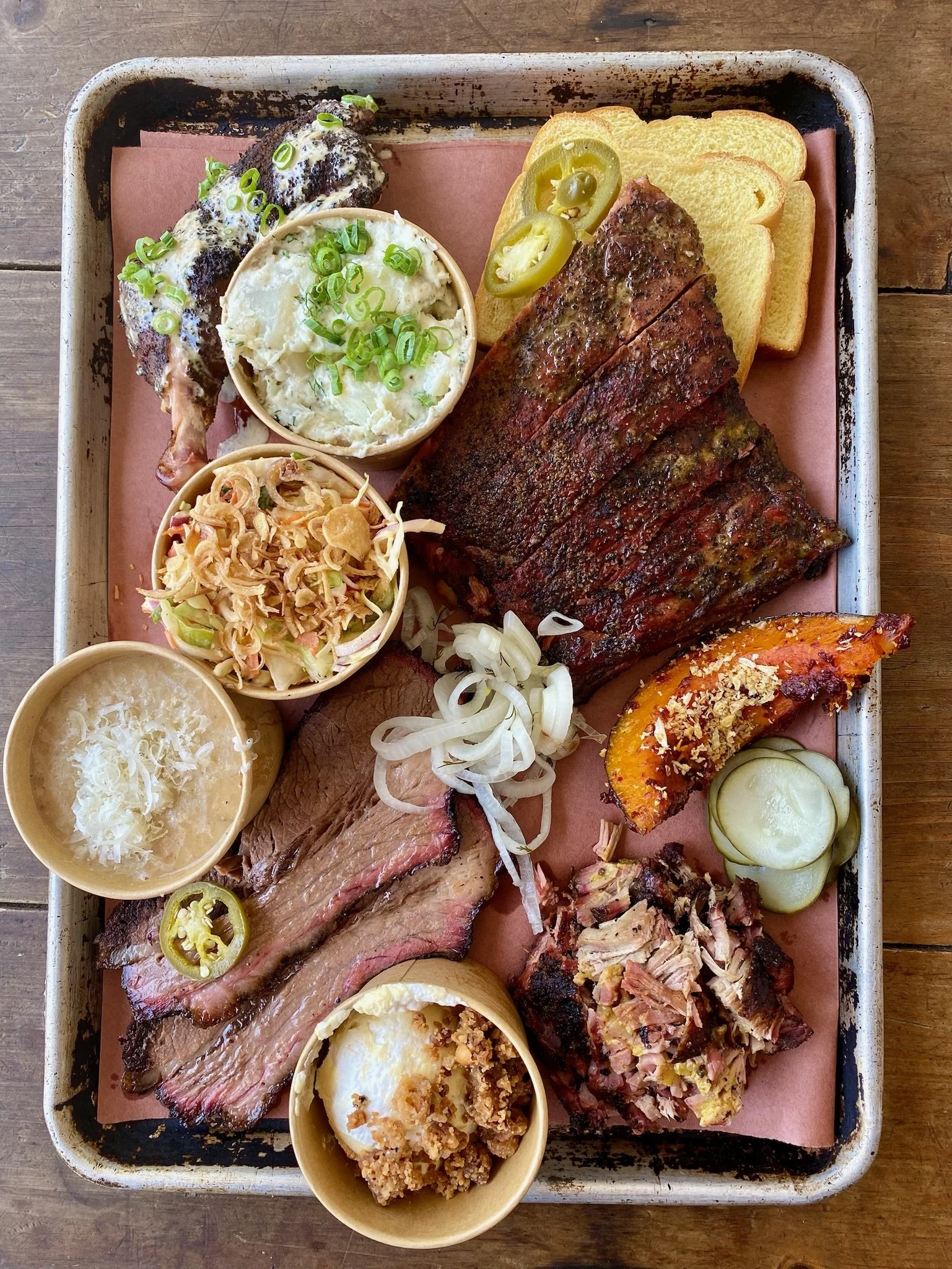 A tray of assorted barbecue meats, sides, and bread, including sliced brisket, ribs, potato salad, cole slaw, baked squash, pickles, and sliced bread with a rustic setting.