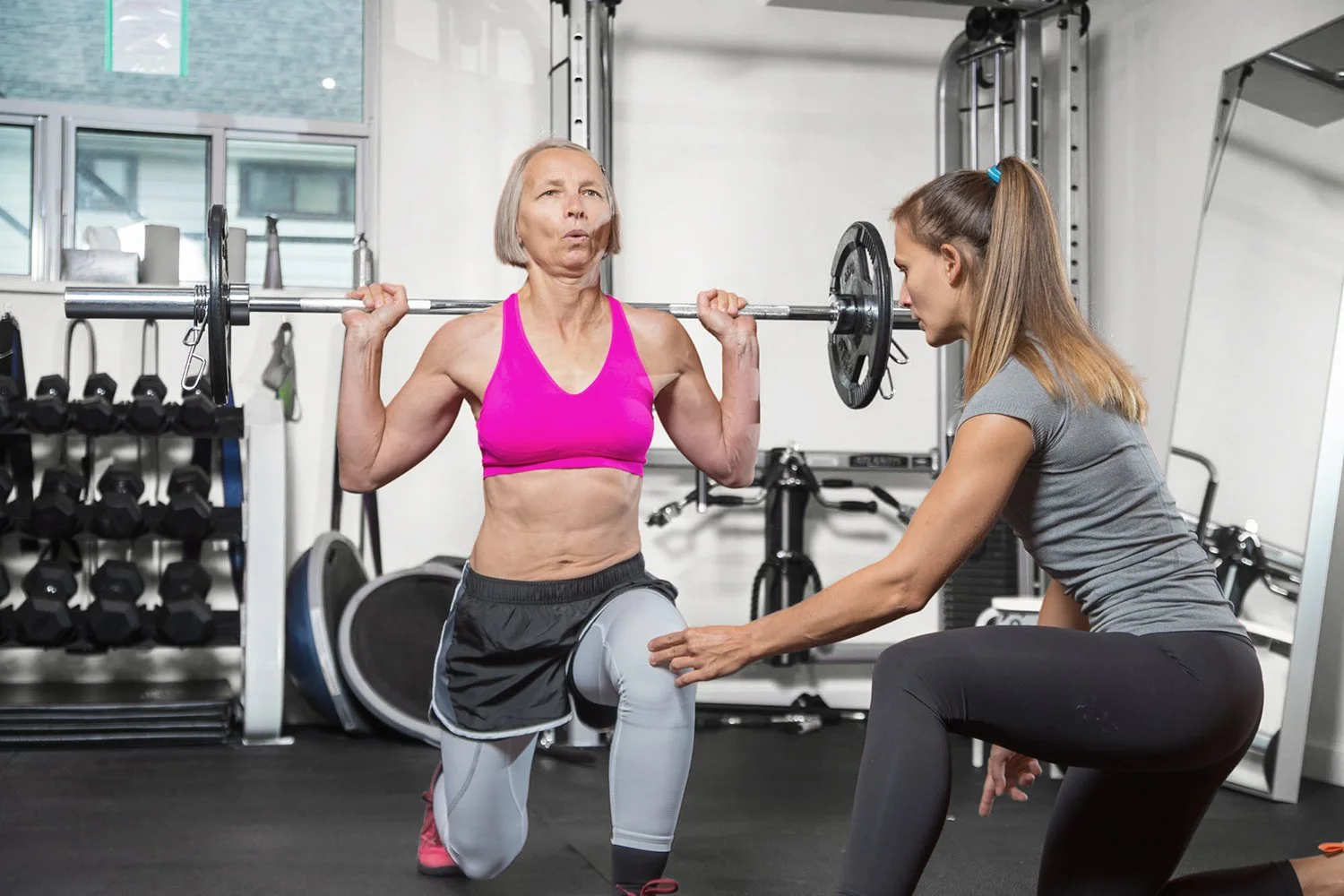 An elderly woman lifting a barbell on her shoulders during a workout, supervised by a young female trainer in a gym.