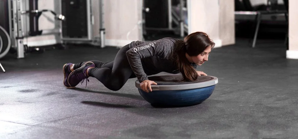 Housefit trainer in workout clothes exercises on a Bosu ball in a gym.