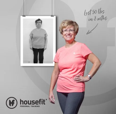 A woman in a pink shirt and gray leggings smiling in front of a photo showing her previous weight loss. The photo has text saying "Lost 50 lbs in 8 months" and the HouseFit Personal Training logo.