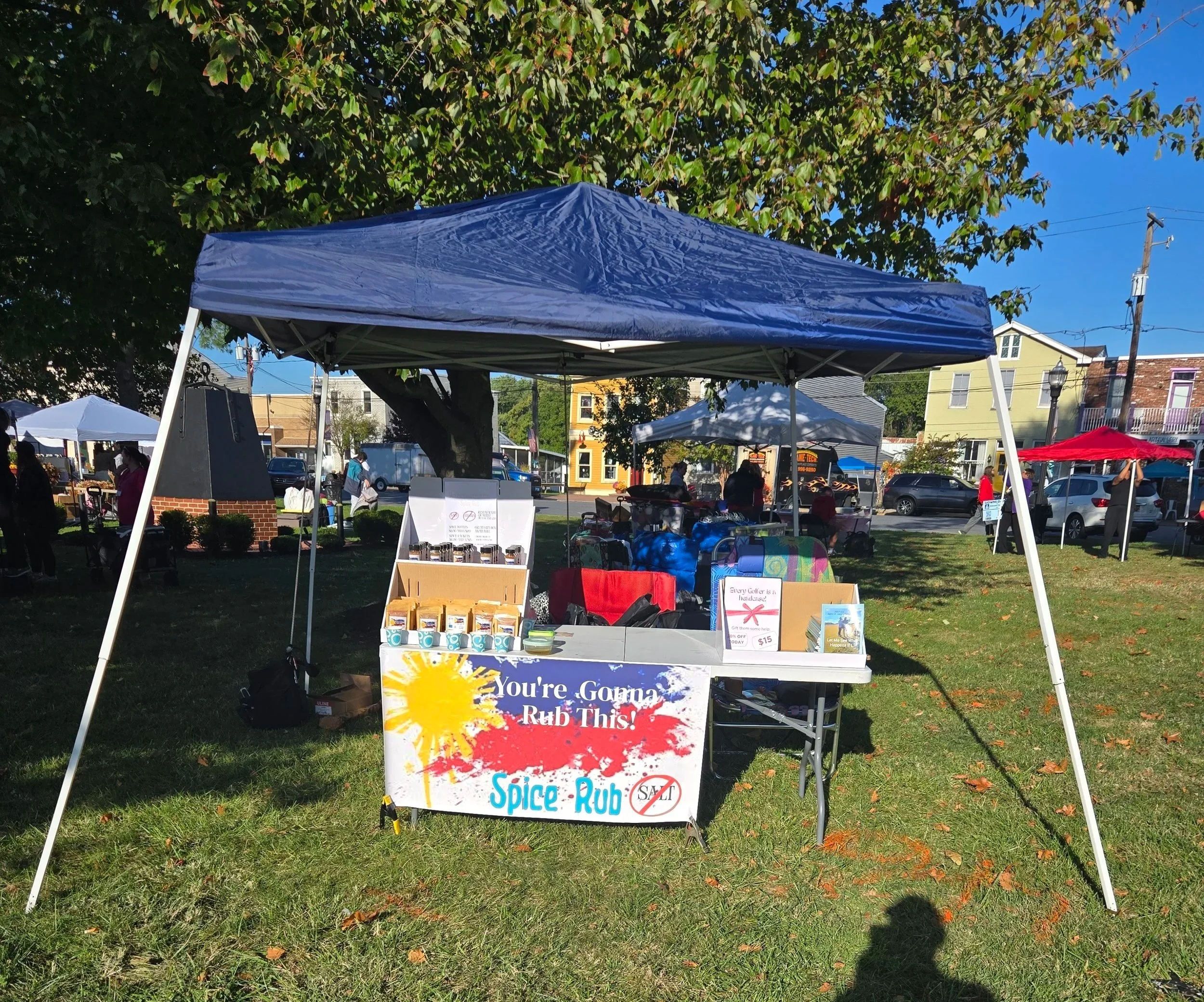 A market stall under a blue canopy with a sign that reads "You're Gonna Rub This NO-SALT Spice Rub" at an outdoor event on a sunny day, with trees, people, and other stalls in the background.