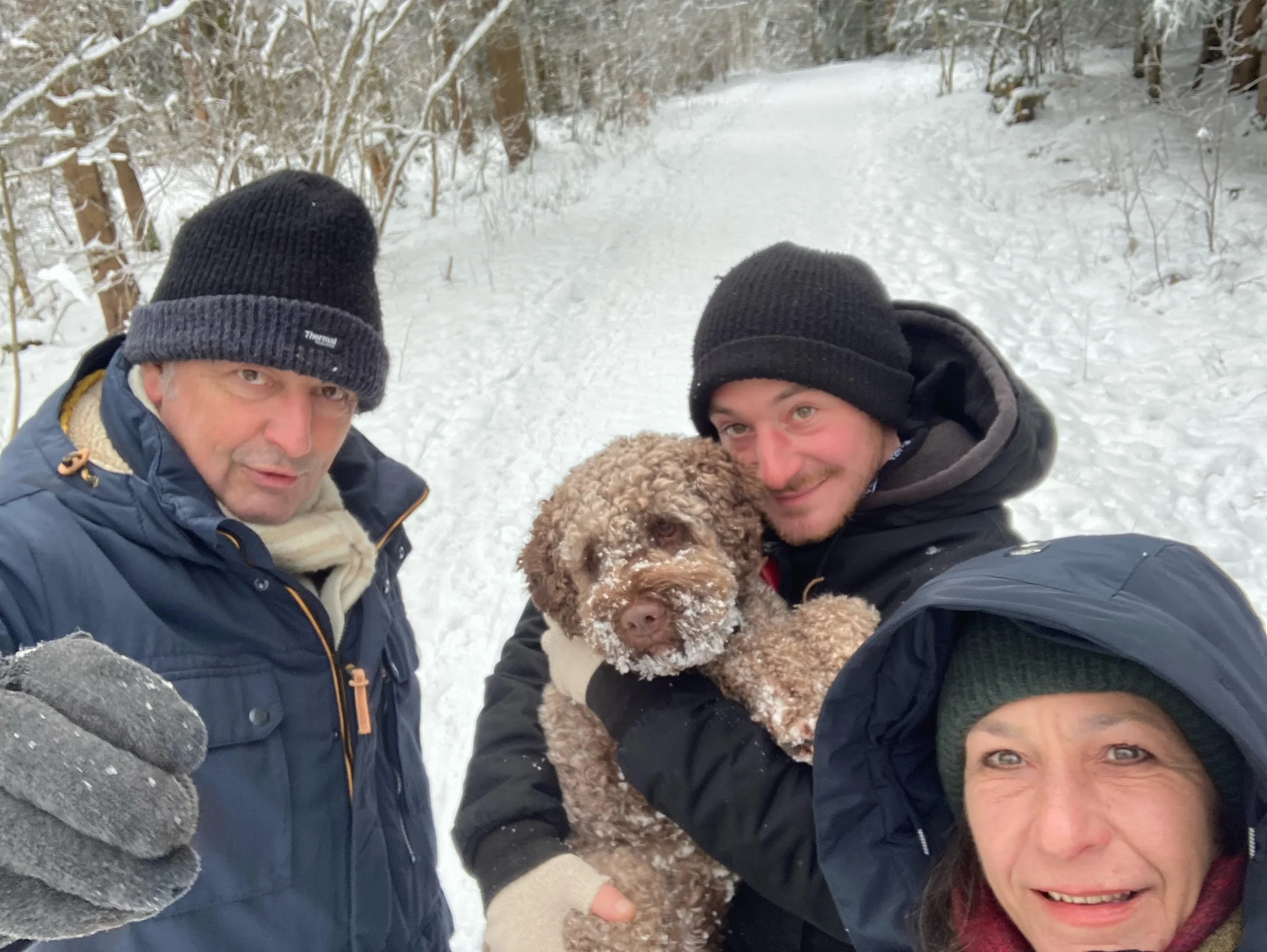 Winzermeister Dieter Amling, David Amling und Edda Amling schauen bei einem Winterspaziergang im Wald gemeinsam mit Trüffelhund Ebbi lachend in die Kamera.