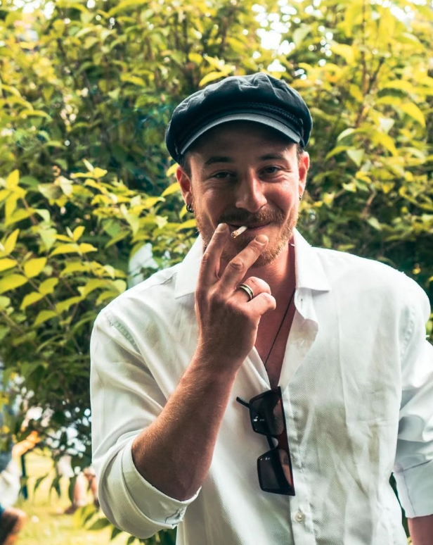 Winzer David Amling mit Hut und weißem Hemd, raucht eine Zigarette, steht vor grünen Büschen und trägt eine schwarze Sonnenbrille um den Hals. Klicke hier, um ihm auf Instagram zu folgen.