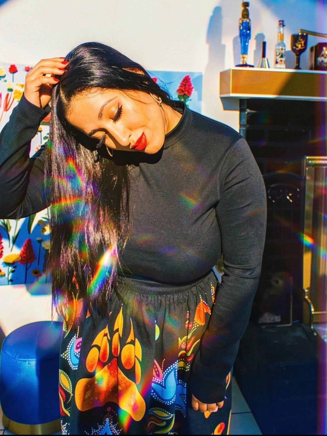 A woman with long dark hair, wearing a black long sleeve shirt and a colorful skirt, stands with her head slightly bent and her hand on her hair. She has bright red lipstick and gold hoop earrings. The background features a colorful floral wallpaper, a wooden shelf with bottles, and a black speaker. There are rainbow light reflections on her and the surroundings.