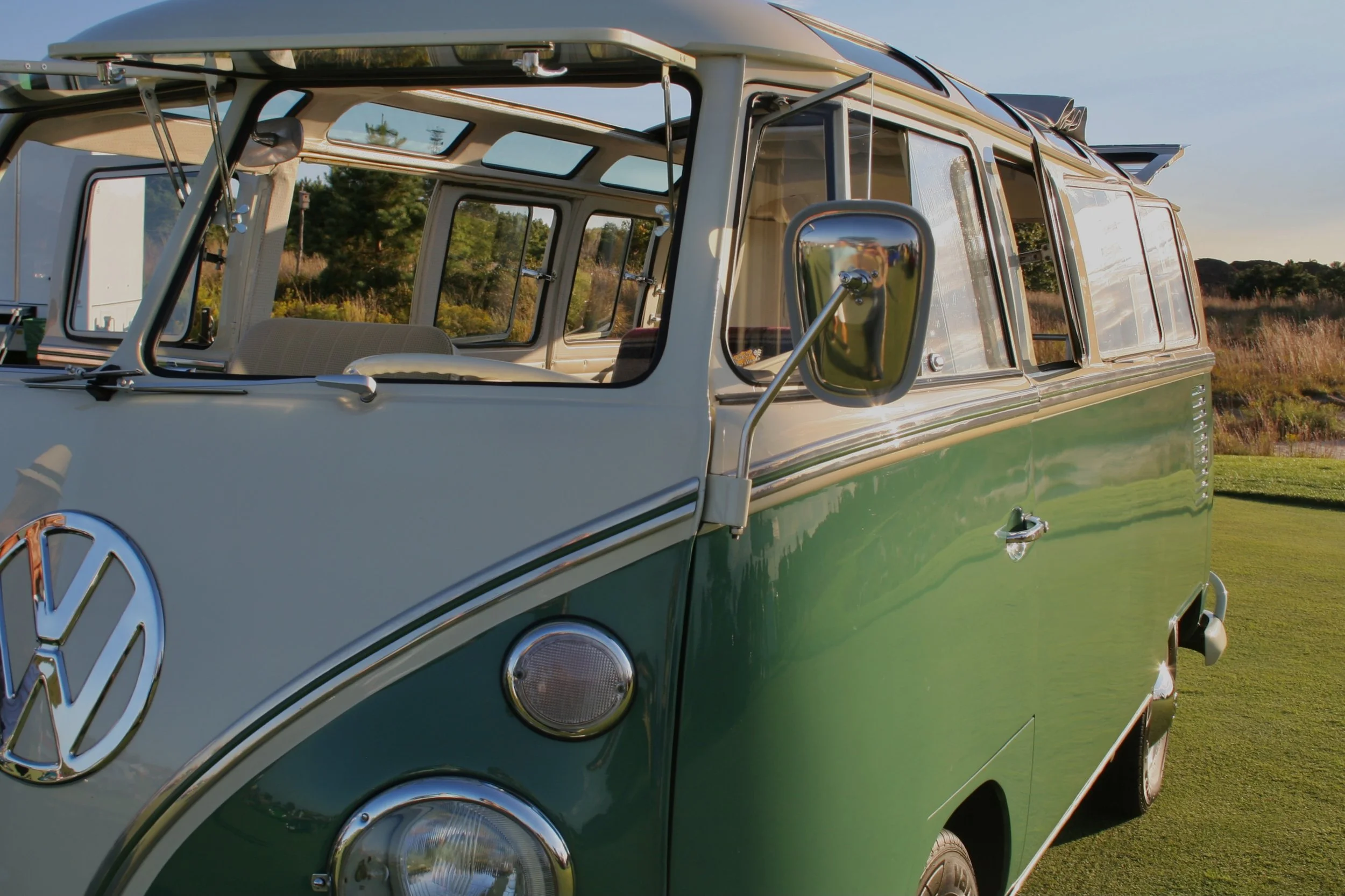 A vintage green and cream Volkswagen Type 2 camper van parked on a grassy field with trees and a clear sky in the background.