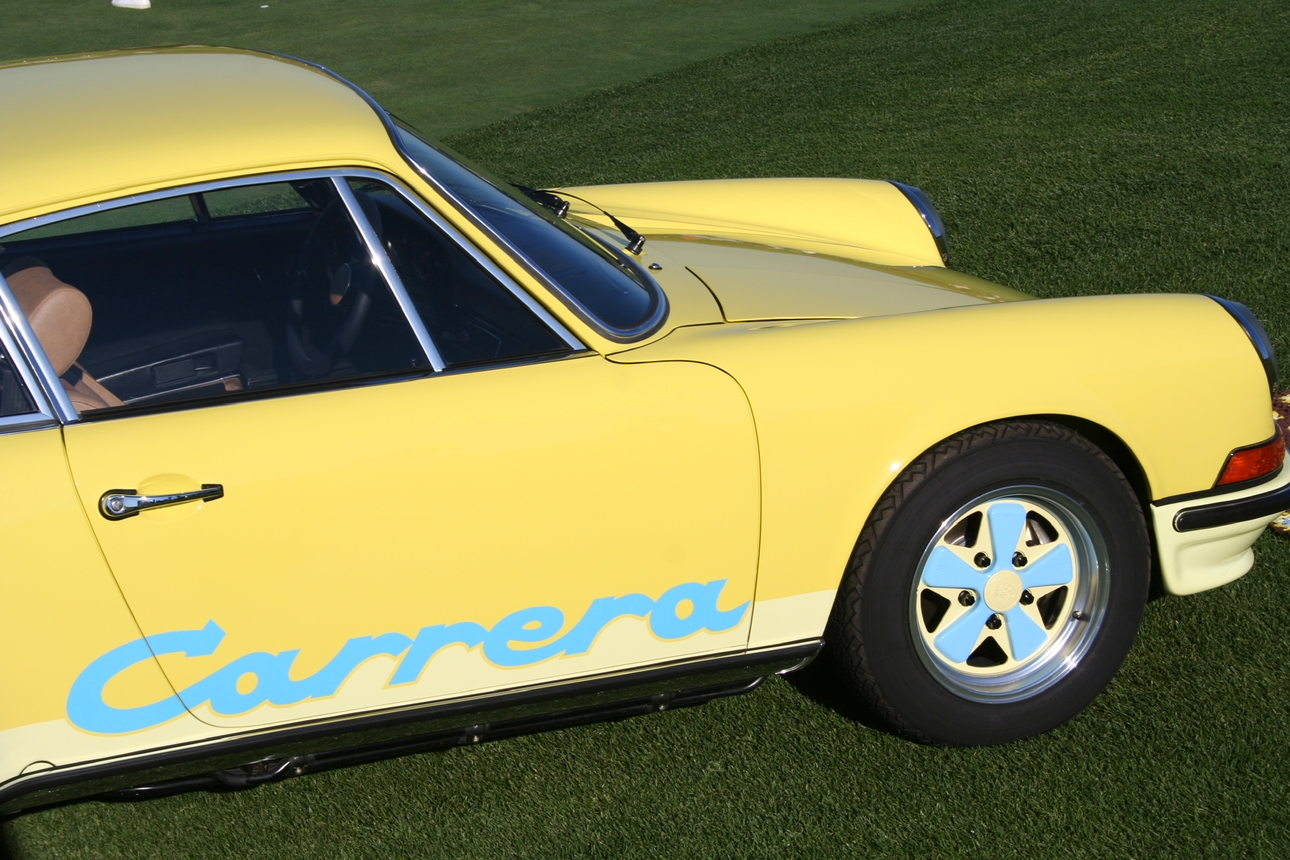 Close-up of a yellow Porsche Carrera sports car parked on green grass, showcasing the front and side, including a part of the wheel and the Carrera logo in blue.