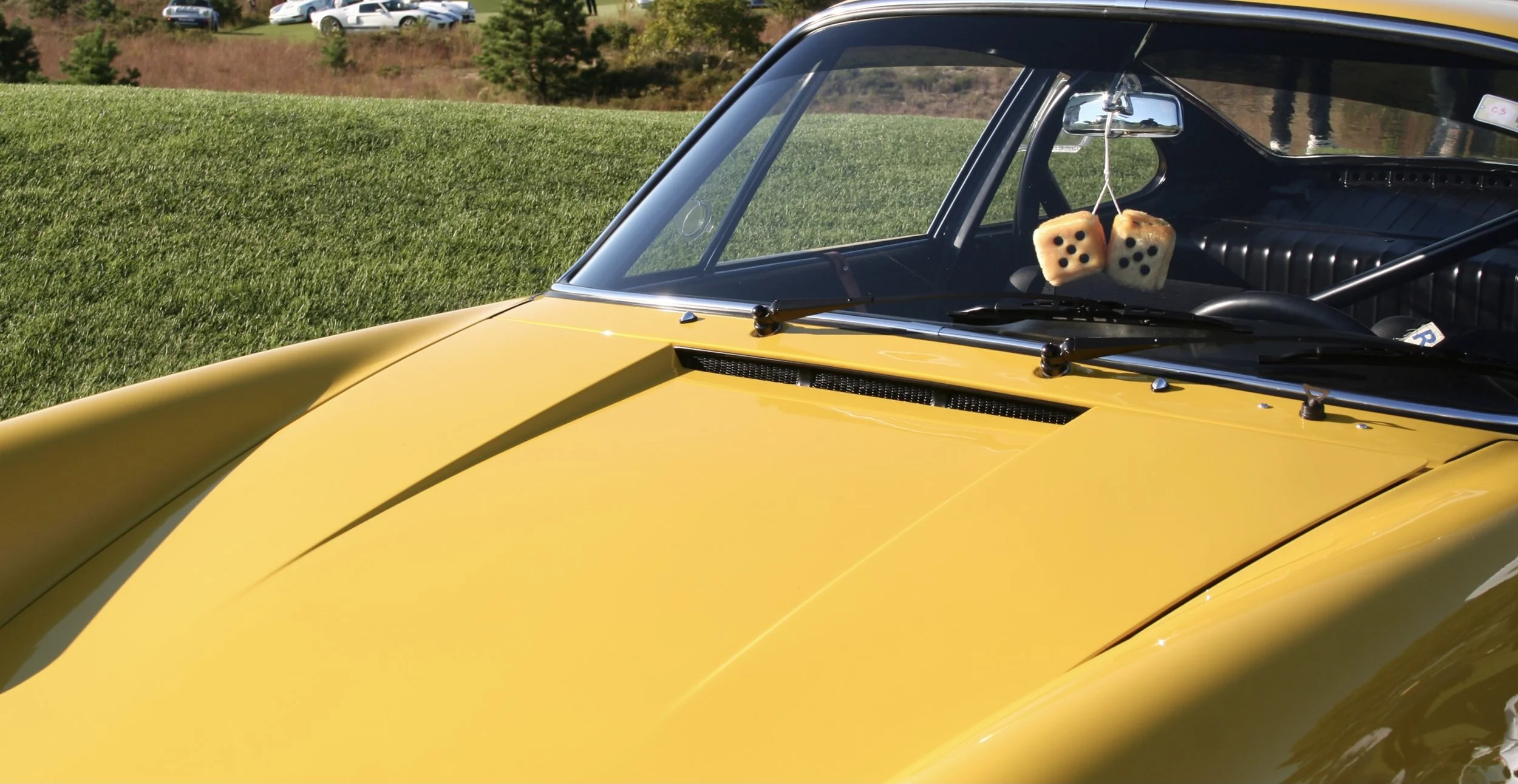 Close-up of a yellow vintage sports car parked on grass, with dice hanging from the rearview mirror inside the car.