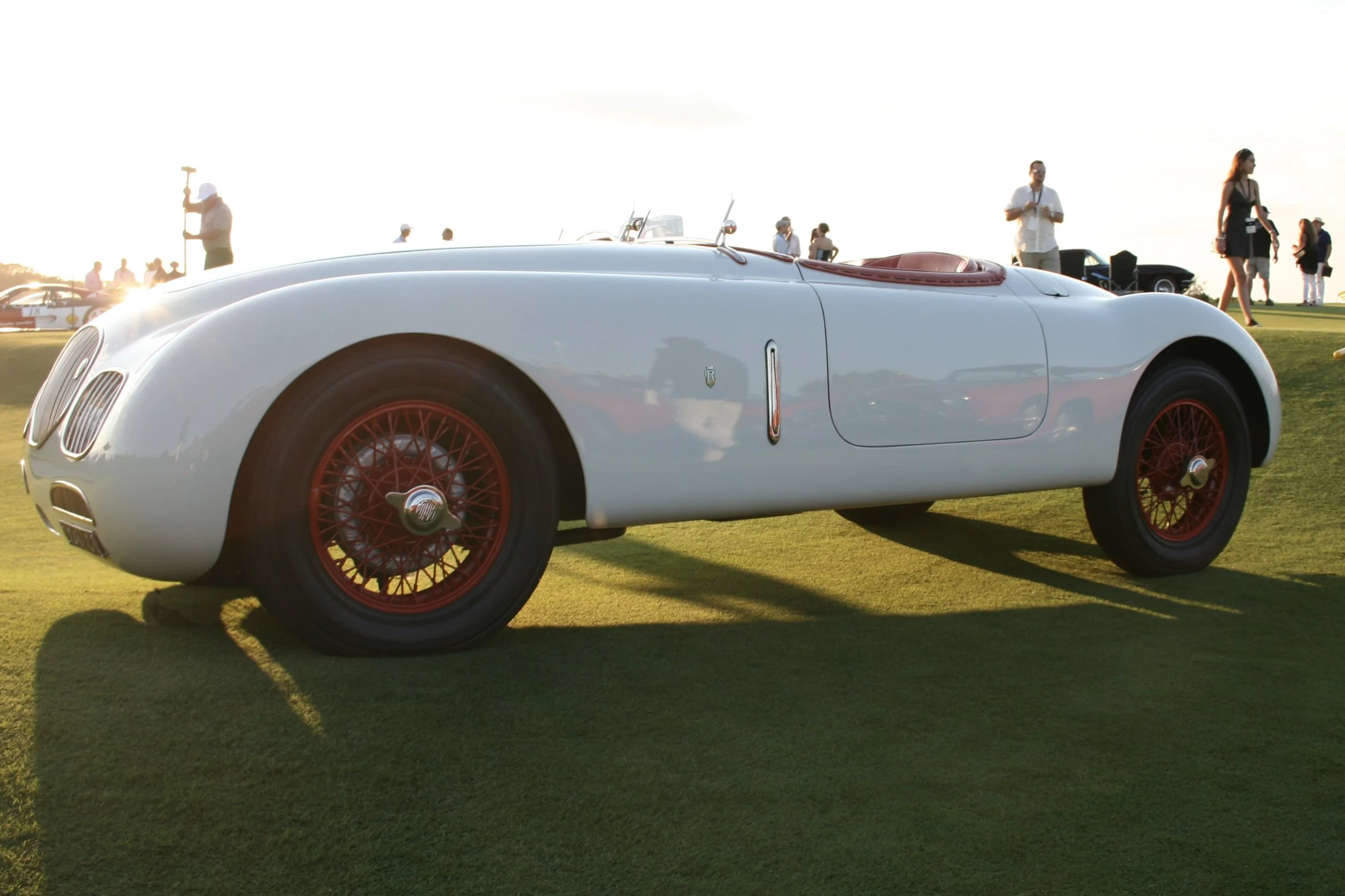 A vintage white sports car with red wire-spoke wheels parked on a grassy area at a car show, with people in the background.