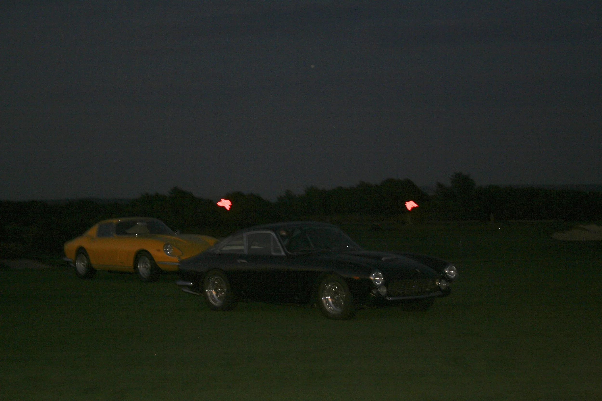 Two vintage cars, a black one and a yellow one, parked on a grassy field at night with a dark sky and faint red lights in the background.