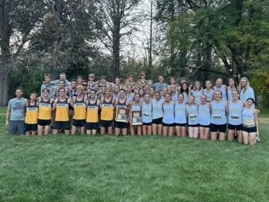 Group of young athletes in athletic uniforms standing outdoors on grass, holding awards, with trees in the background.