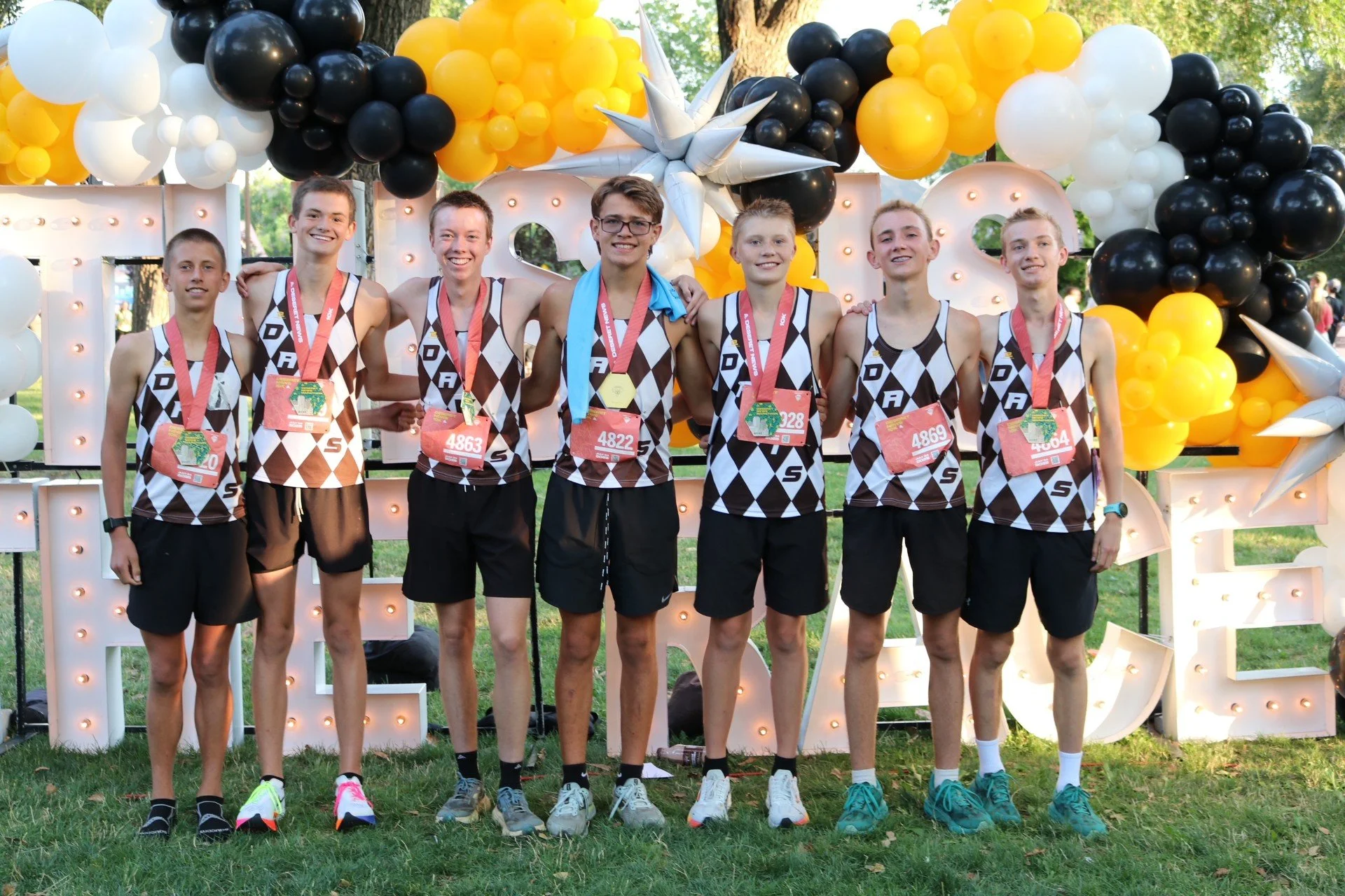 Seven young male athletes wearing similar black and white checkered uniforms stand side by side, each with a medal around their neck, in front of a decorated backdrop of black, white, and yellow balloons. They are outdoors on grass and are smiling at