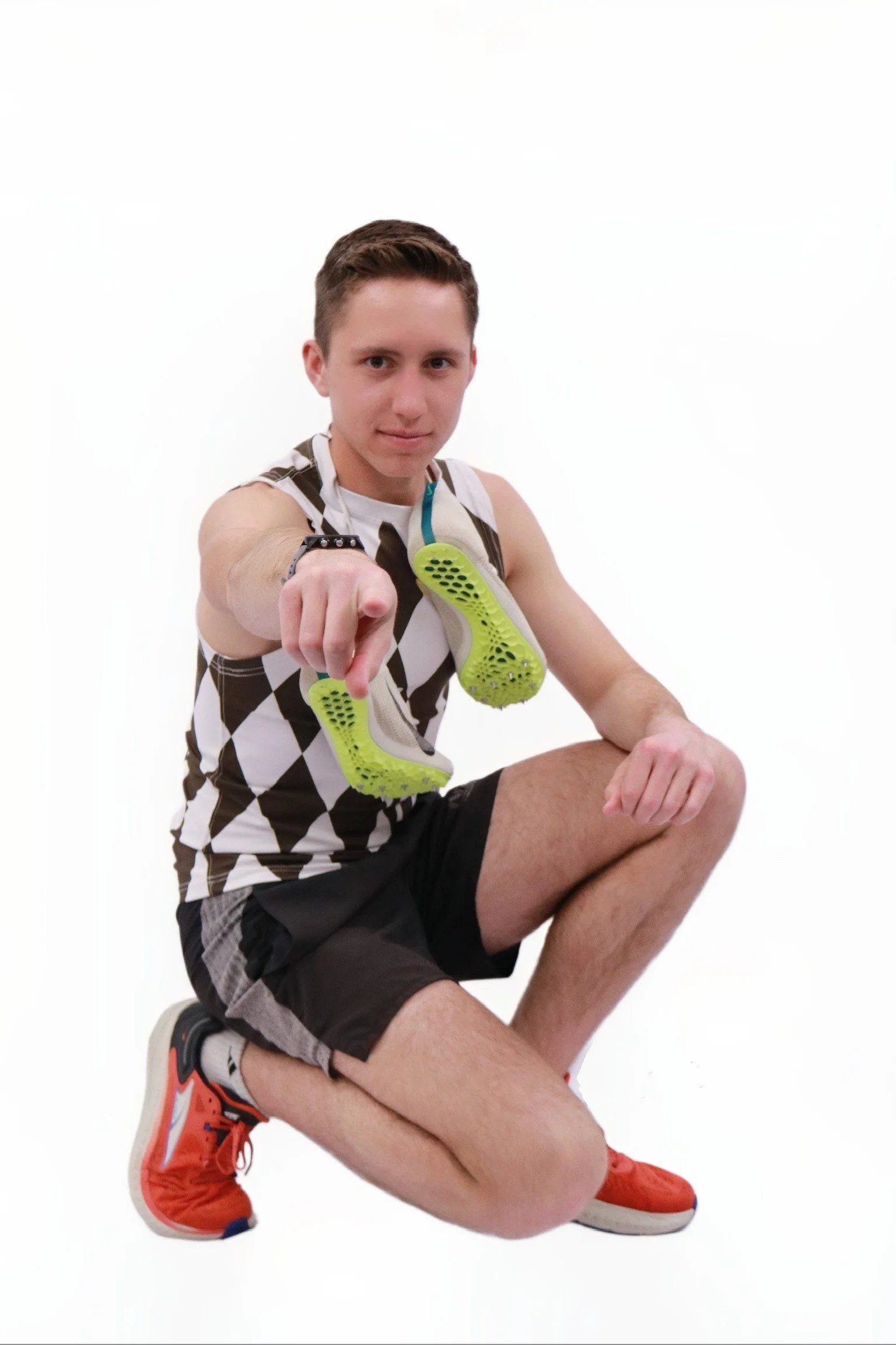 Young man crouching down, holding a pair of lime green running shoes, wearing a black and white checkered sleeveless shirt, black shorts, orange running shoes, and a fitness tracker, with a confident expression on a white background.