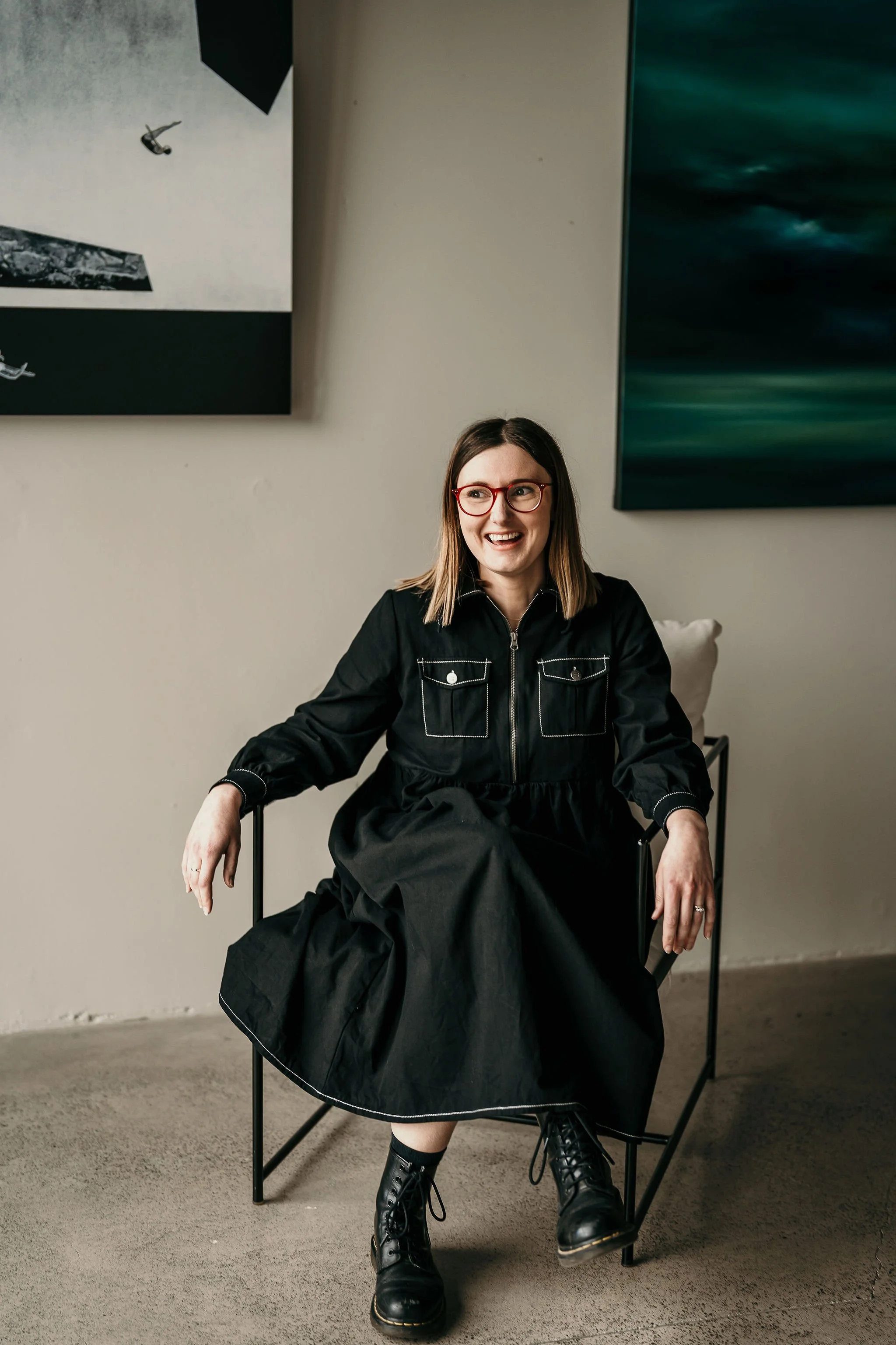 Woman with shoulder-length brown hair, wearing glasses, a black dress, and black combat boots, sitting on a chair in an art gallery with abstract paintings on the wall.