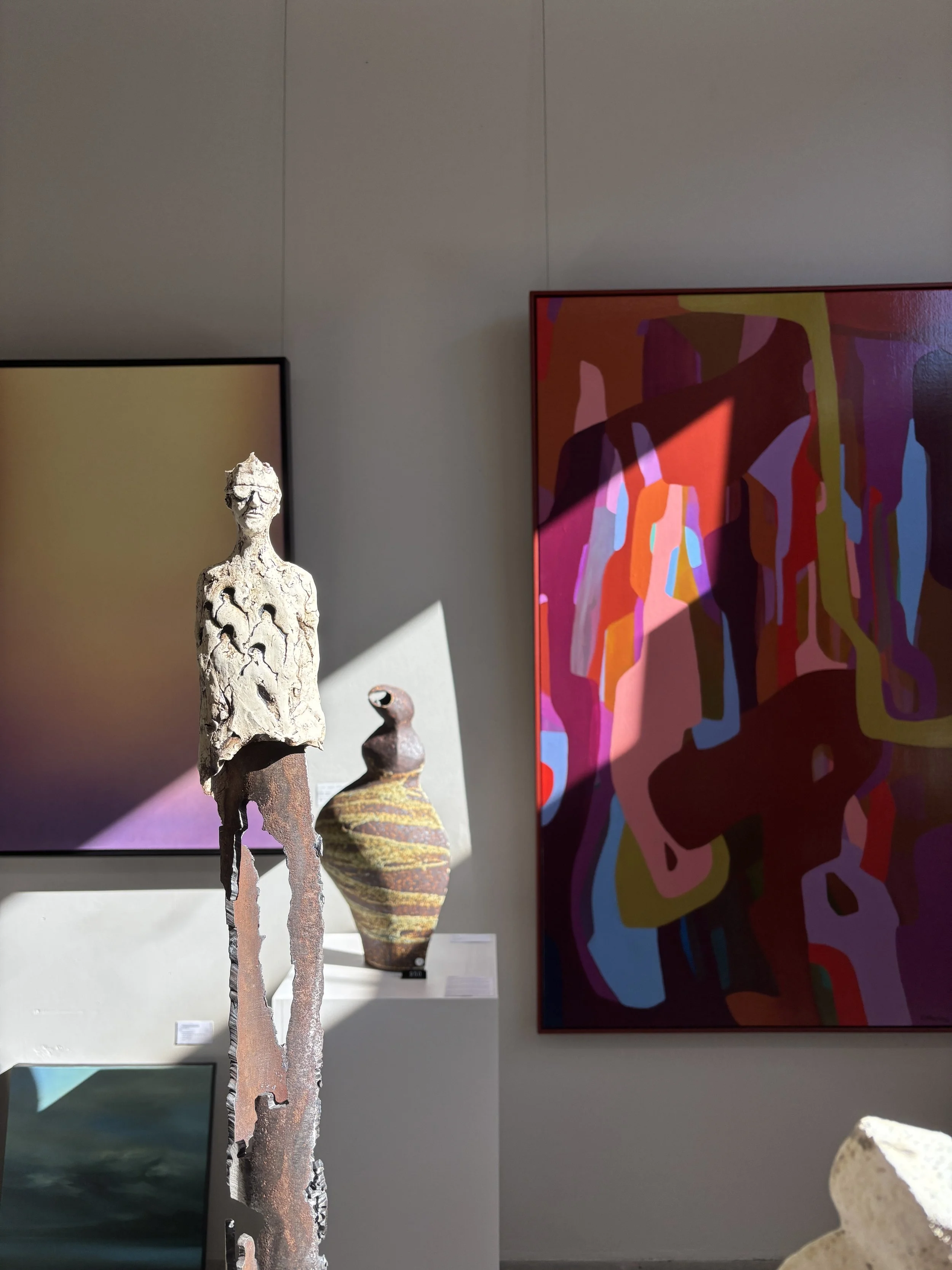 Art gallery interior with an abstract multicolored painting, sculptural human figure, framed artwork, and decorative pottery.