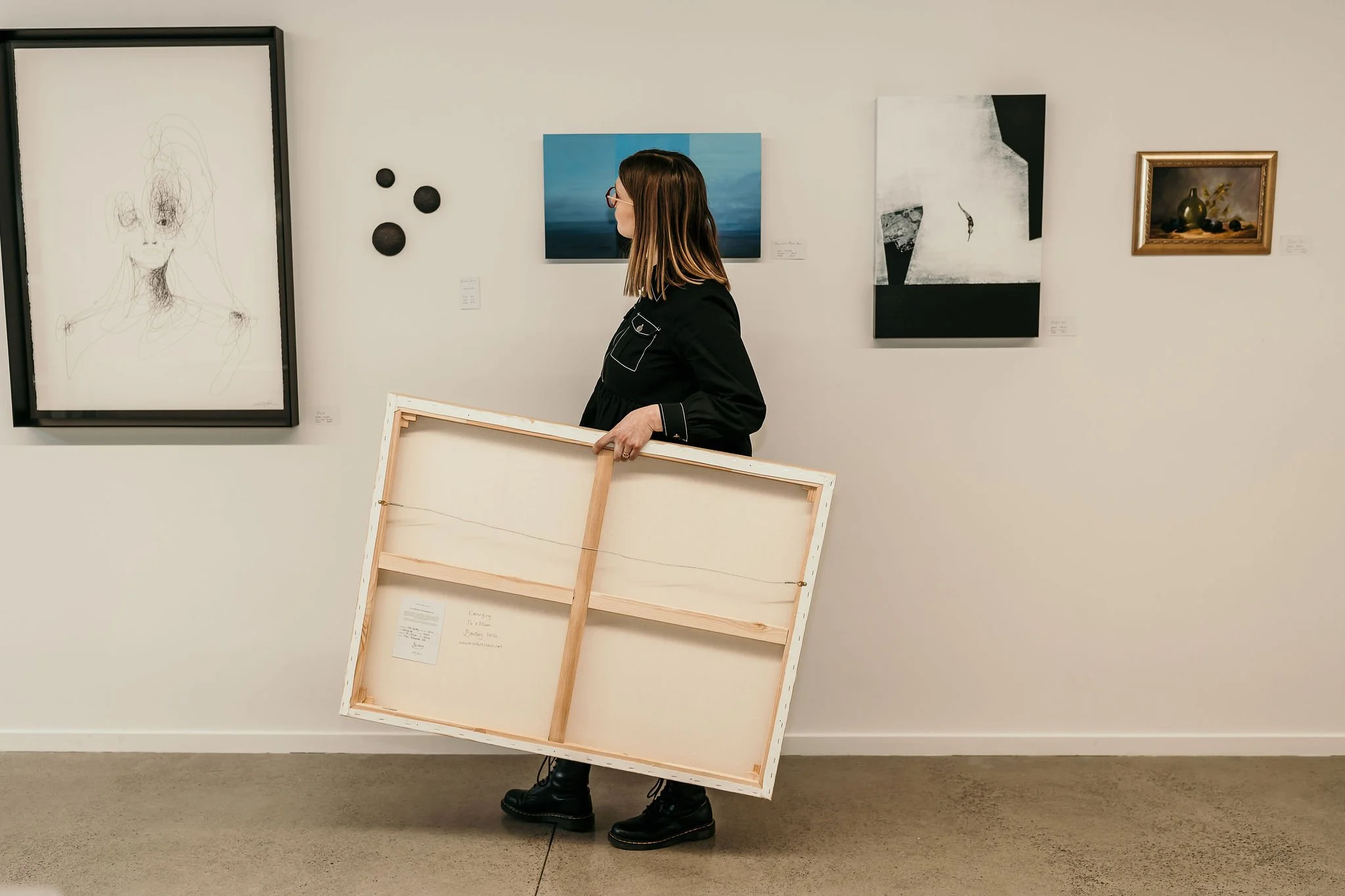 Woman walking through an art gallery holding a large painted canvas viewing artworks hung on the wall.