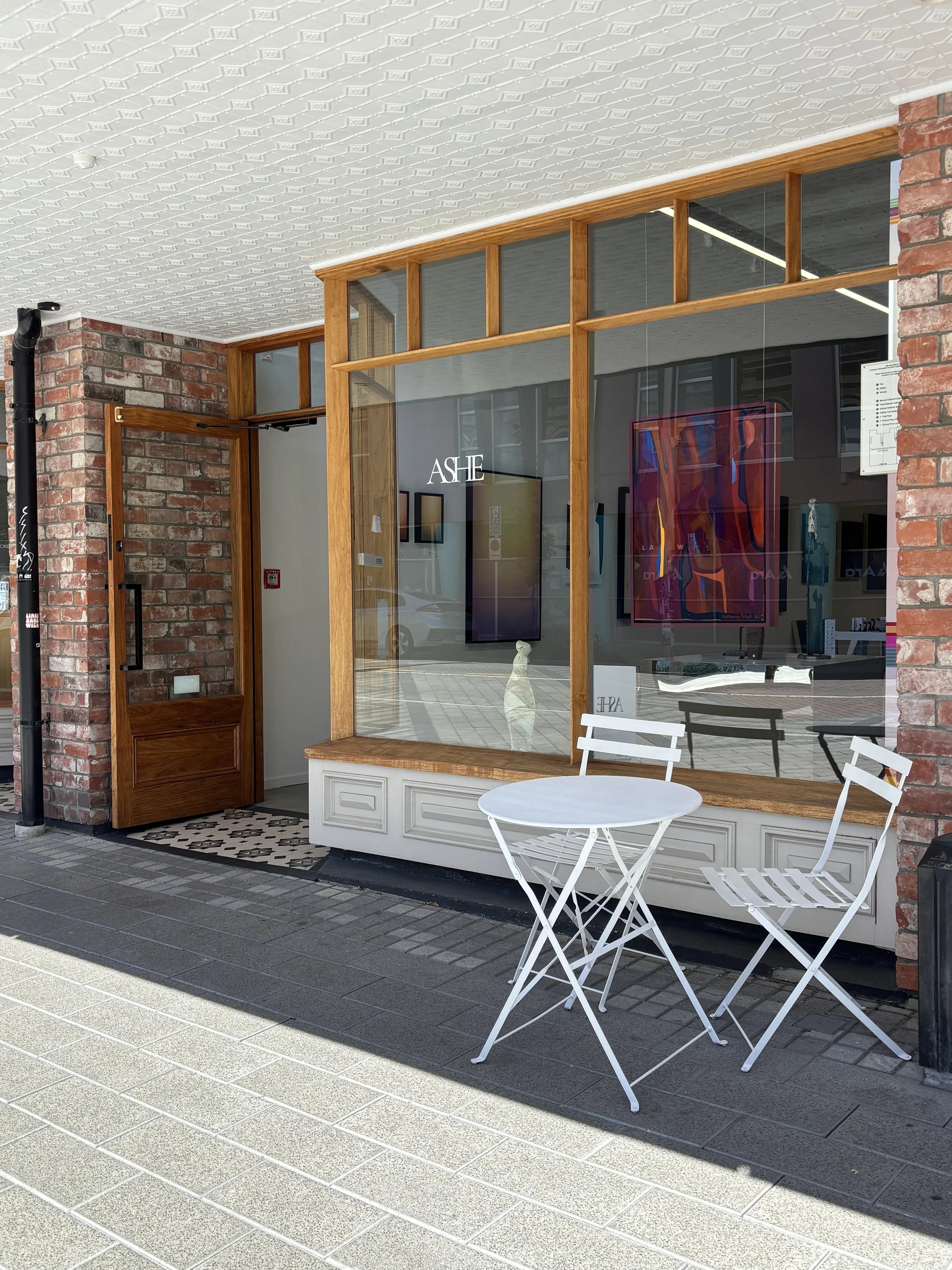 Street-level view of an art gallery storefront with brick walls, wood-framed windows, artwork displays, and white outdoor café furniture.