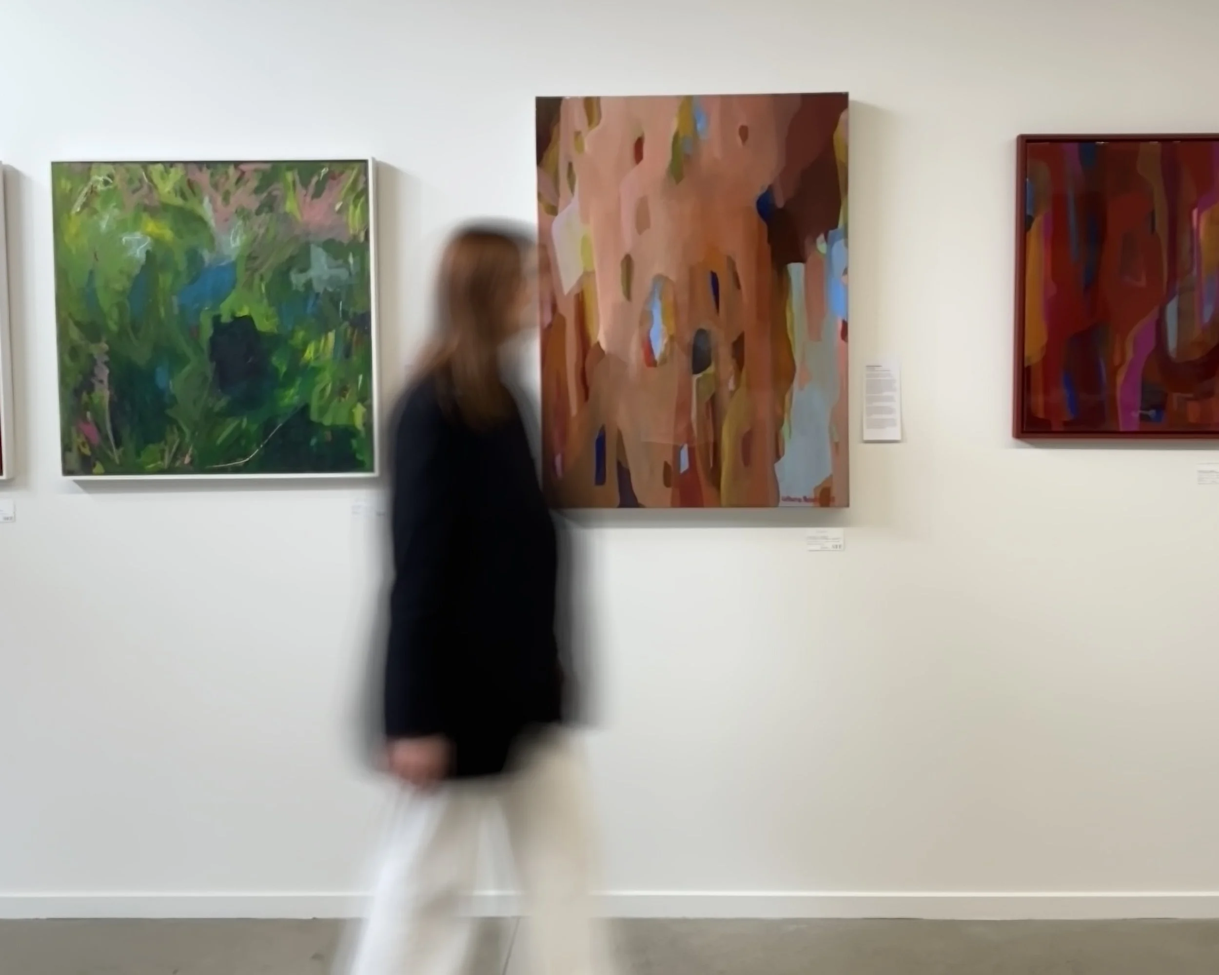 Blurred image of an art gallery with paintings on the wall and a woman with long brown hair walking past.