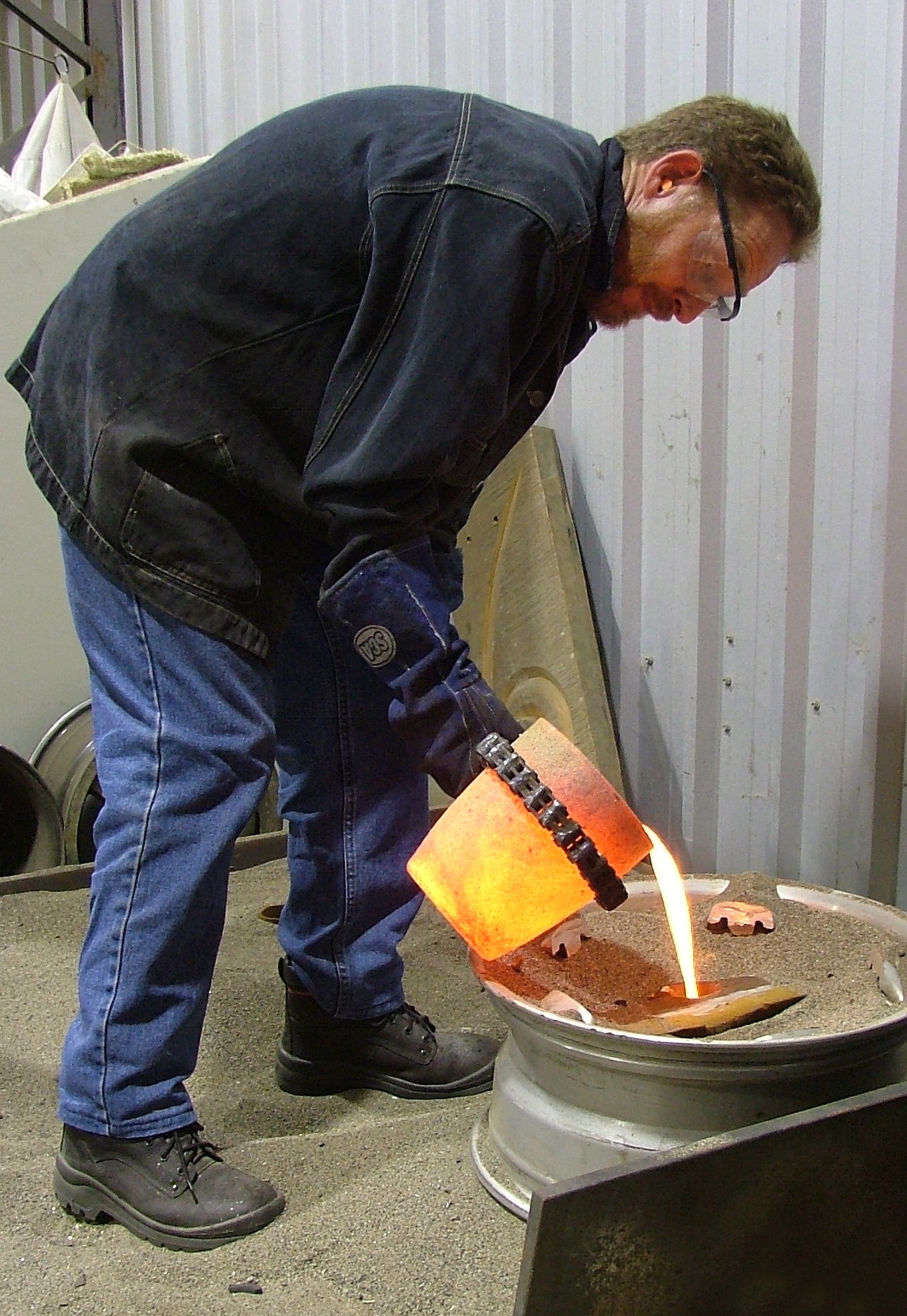 A man in black jeans, black boots, and a black jacket is pouring molten metal into a mold in an industrial workshop.