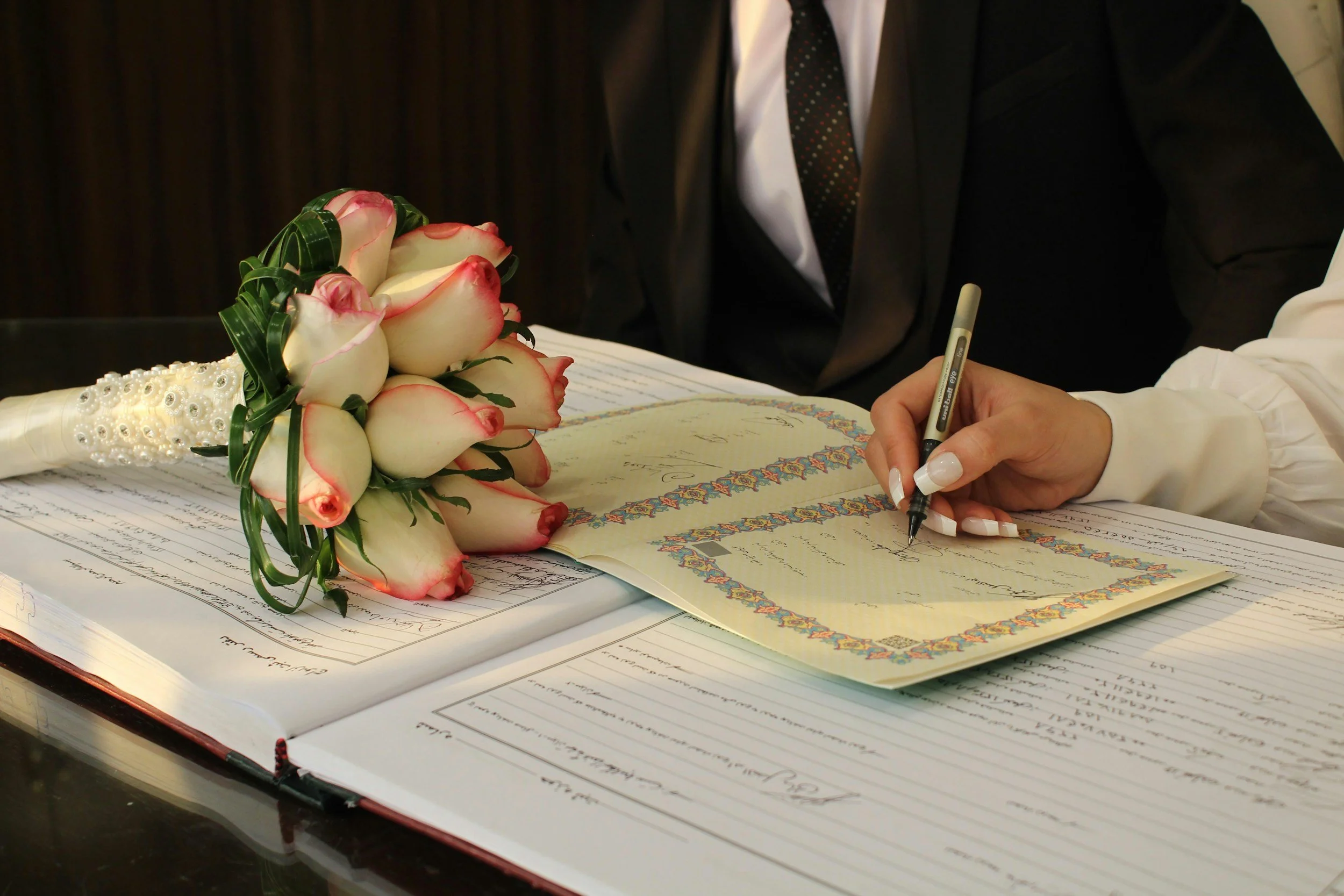 Person signing a marriage register during a wedding ceremony with a bride's bouquet of white roses with pink edges on the table.