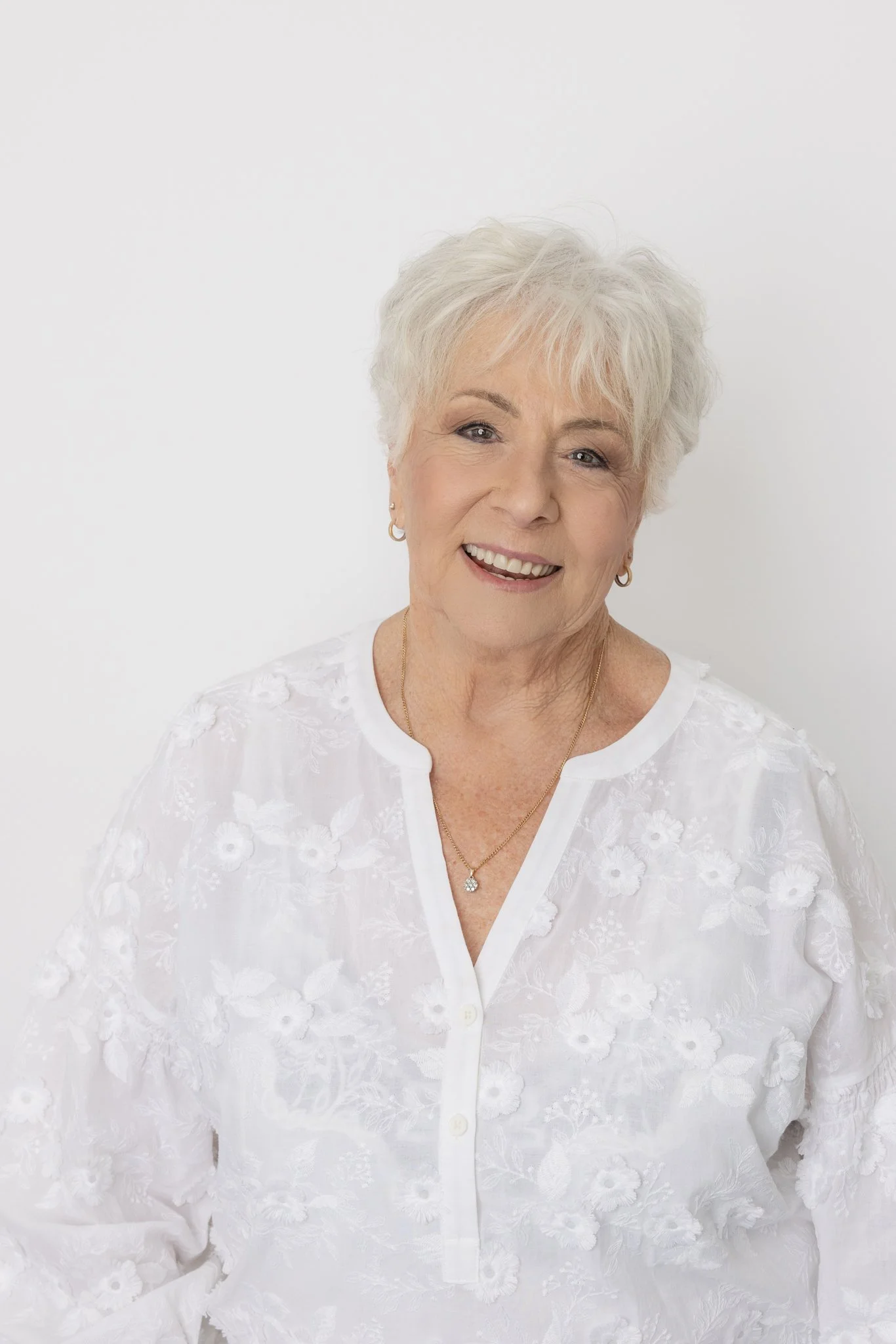 Smiling elderly woman with short white hair wearing a white embroidered blouse and gold jewelry, standing against a plain white background.